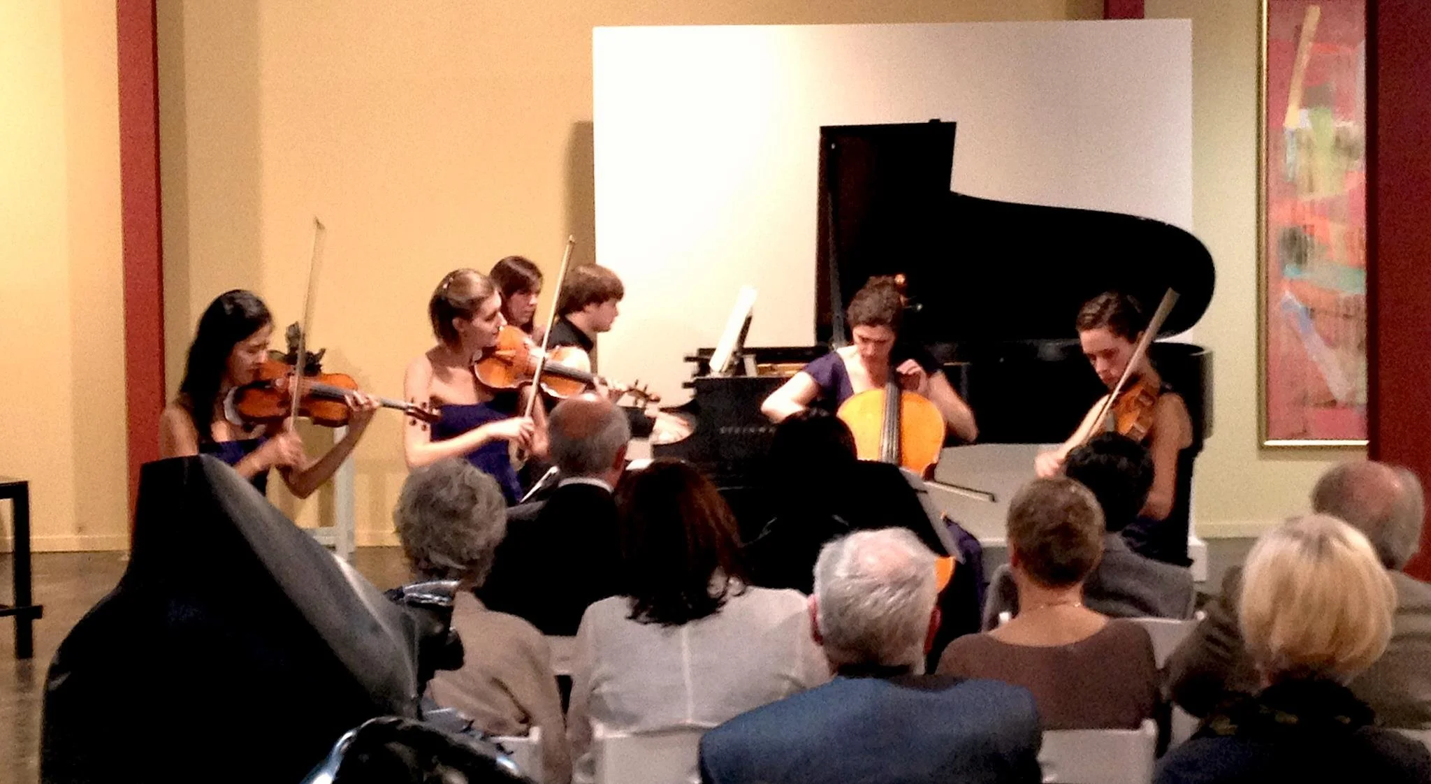 A string quartet performing on stage with a grand piano in the background, audience seated in front watching.