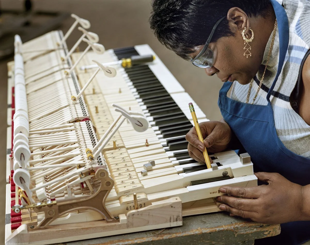 A woman working on the inside of a piano, repairing or tuning it, with a pencil in her hand and wearing safety glasses and a blue apron.