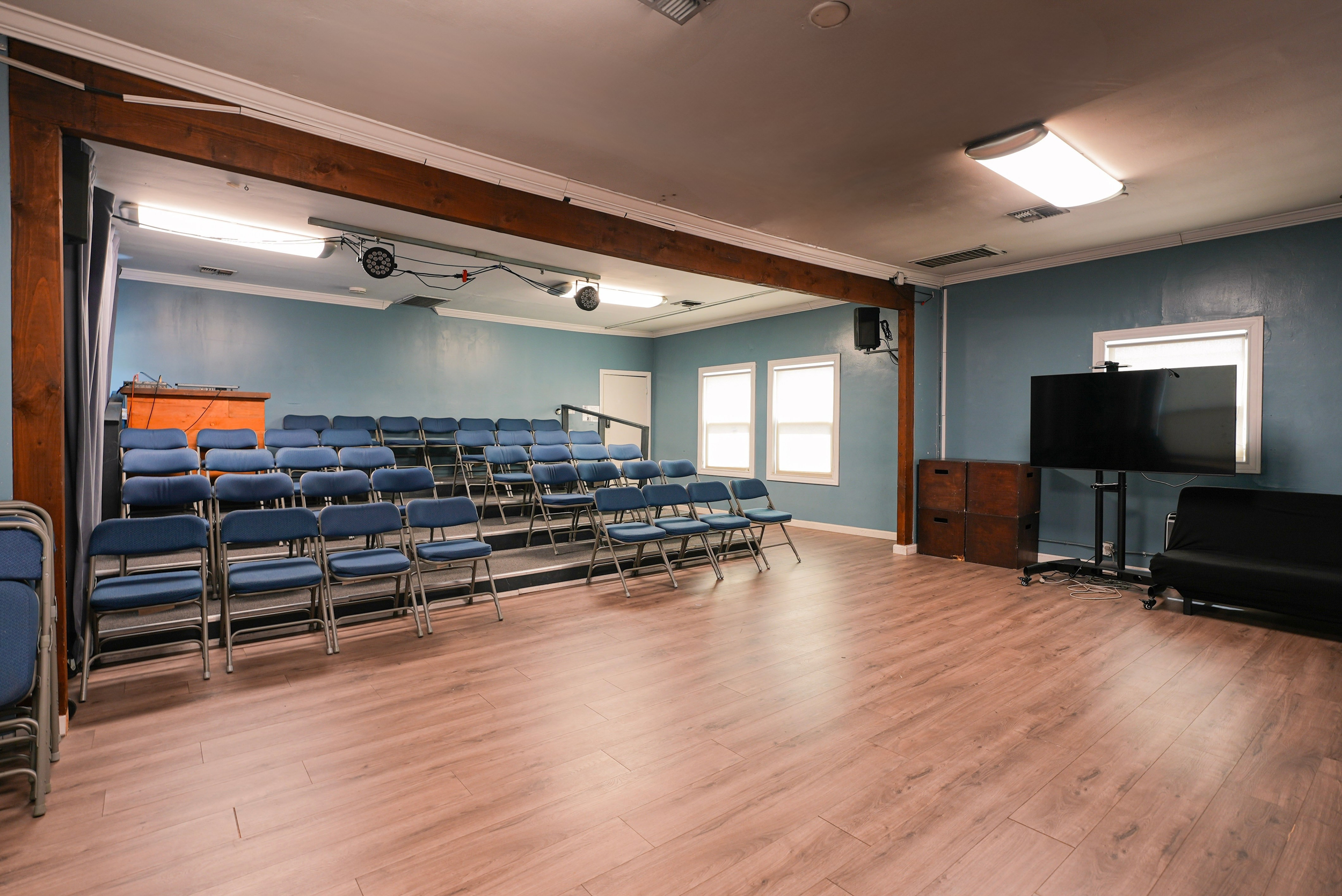 Studio C at Lounge on Melrose Empty blue-walled acting studio with tiered blue chairs, stage lighting, a podium, large display screen, and wood flooring
