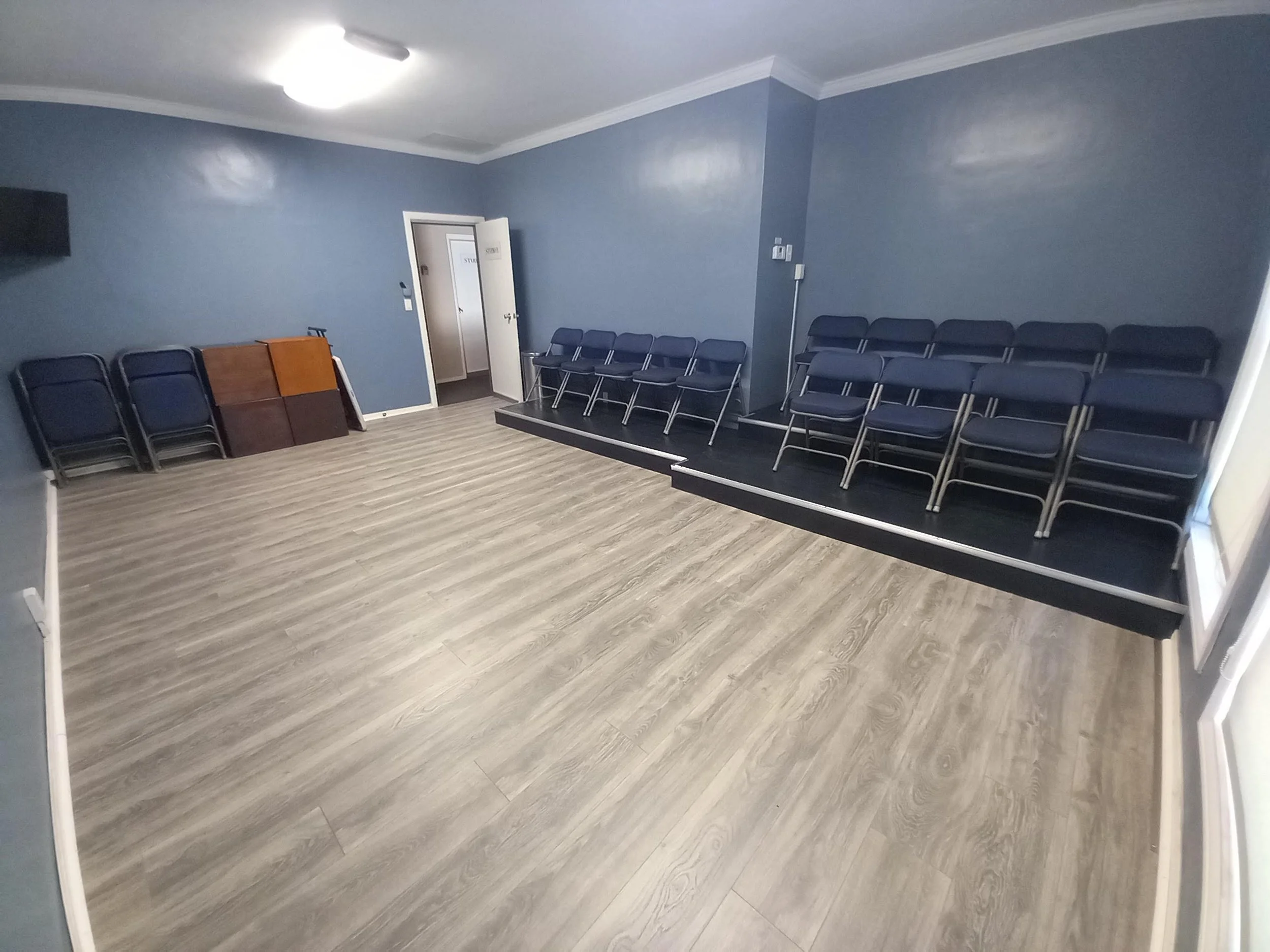 Studio B at Lounge on Melrose Empty blue-walled acting studio with rows of folding chairs, a small raised platform, podium, wood-look flooring, and ceiling light.