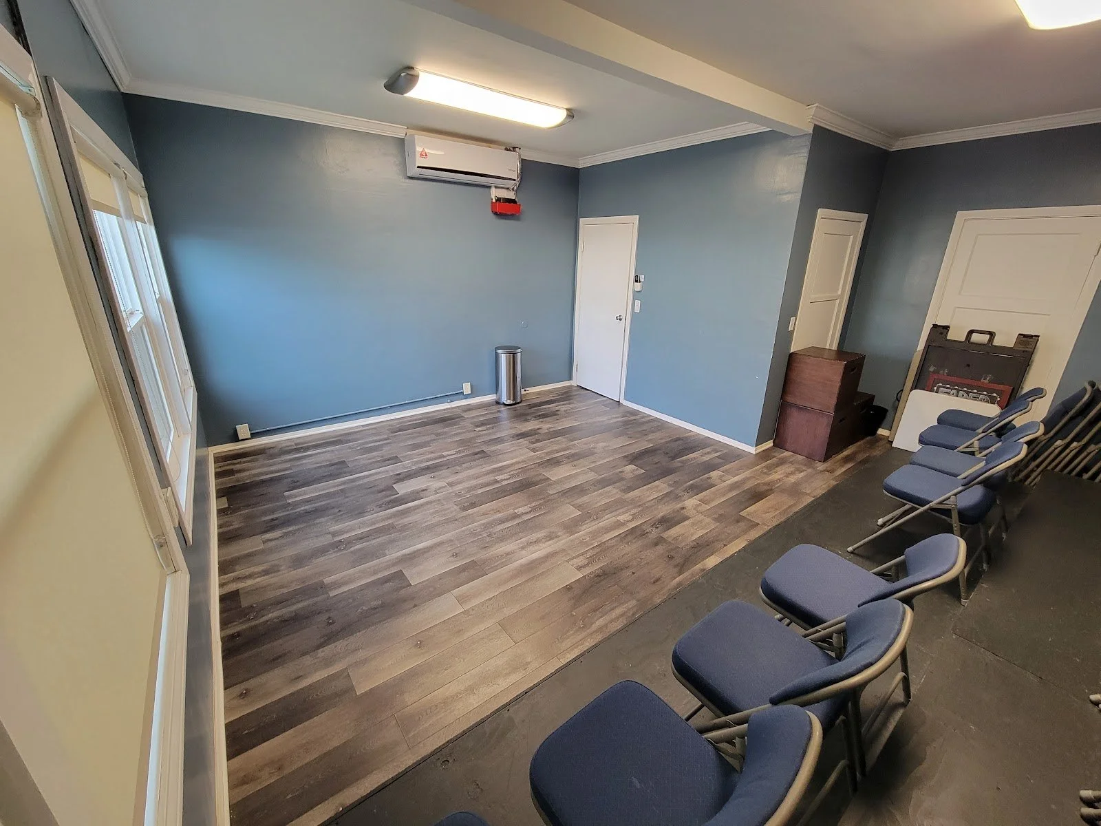 Studio A at Lounge on Melrose Empty blue-walled acting studio with wood-look flooring, folding chairs, doors, windows, air conditioner, and overhead fluorescent lights