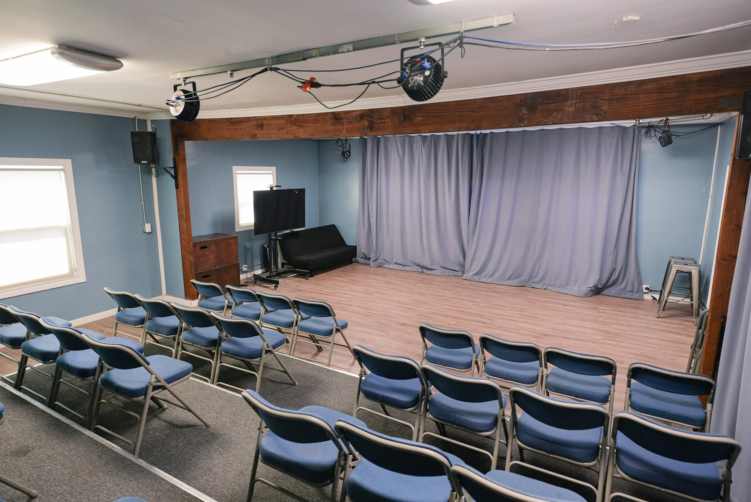 Studio D at Lounge on Melrose Small event venue with a curtained stage, blue folding chairs, speakers, and stage lighting