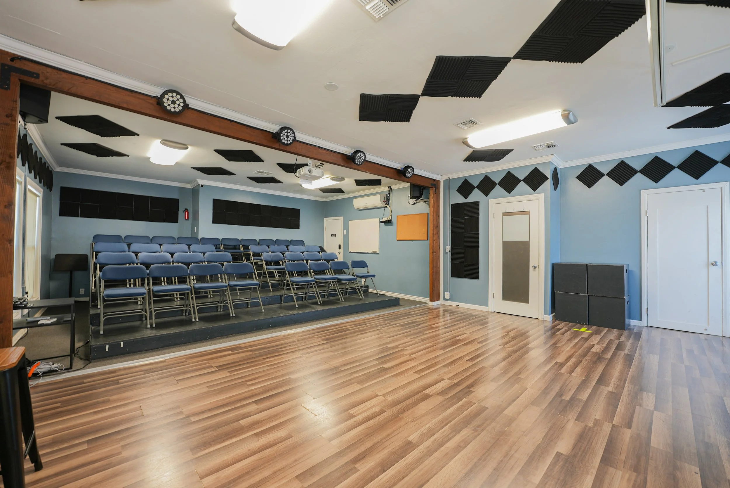 Studio E at Lounge on Melrose Blue-walled acting studio  with tiered rows of folding chairs, acoustic foam panels, stage lighting, projector, and polished wood floor