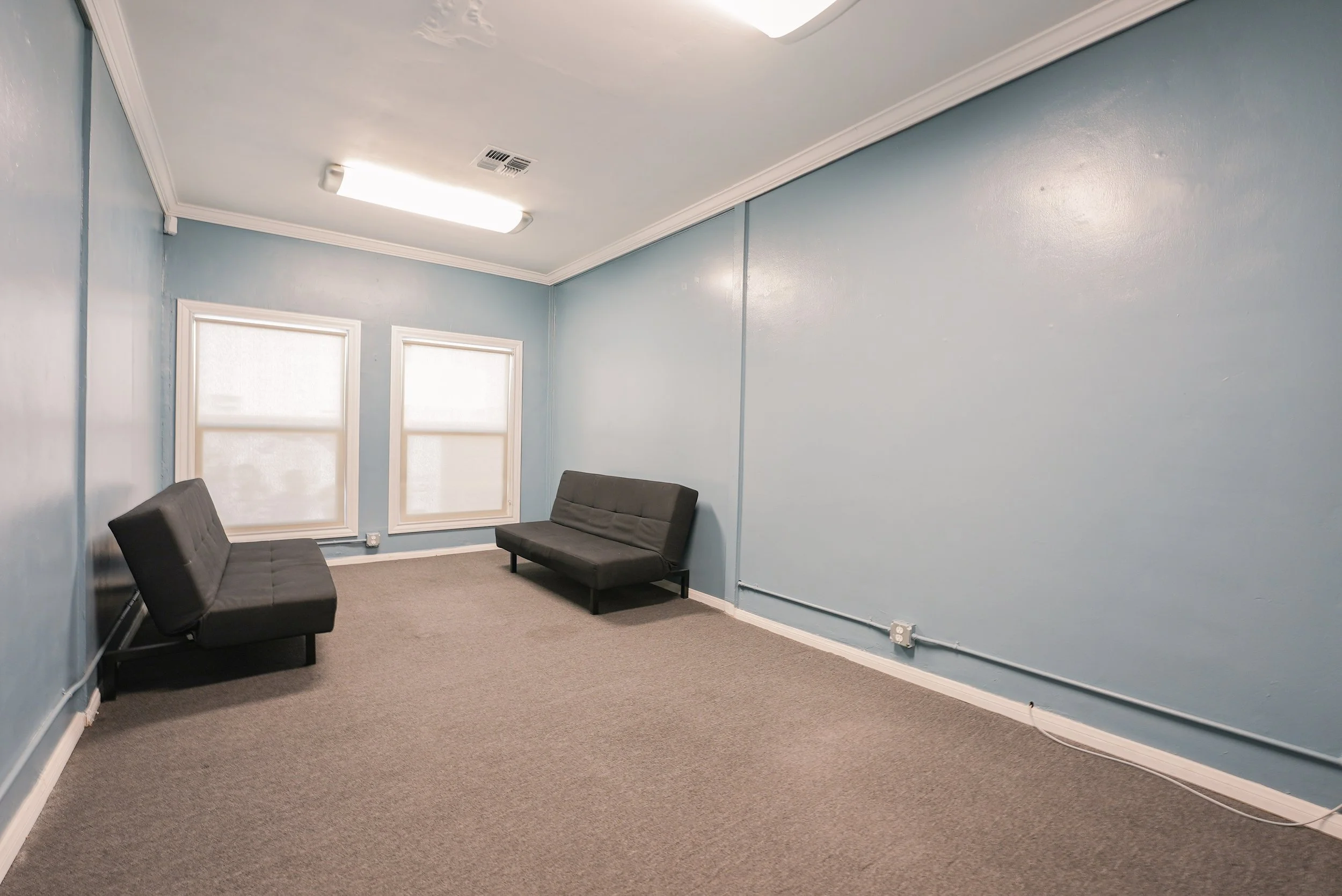 Studio F at Lounge on Melrose Empty blue-painted room with two black futons, carpeted floor, two windows, and fluorescent ceiling lights