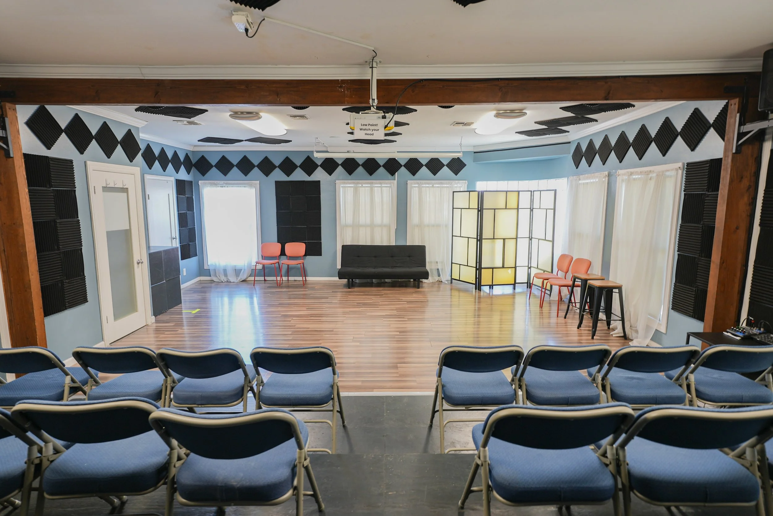 Studio E at Lounge on Melrose rehearsal or performance room with rows of blue chairs, black acoustic foam panels, a couch, stools, and stage equipment.