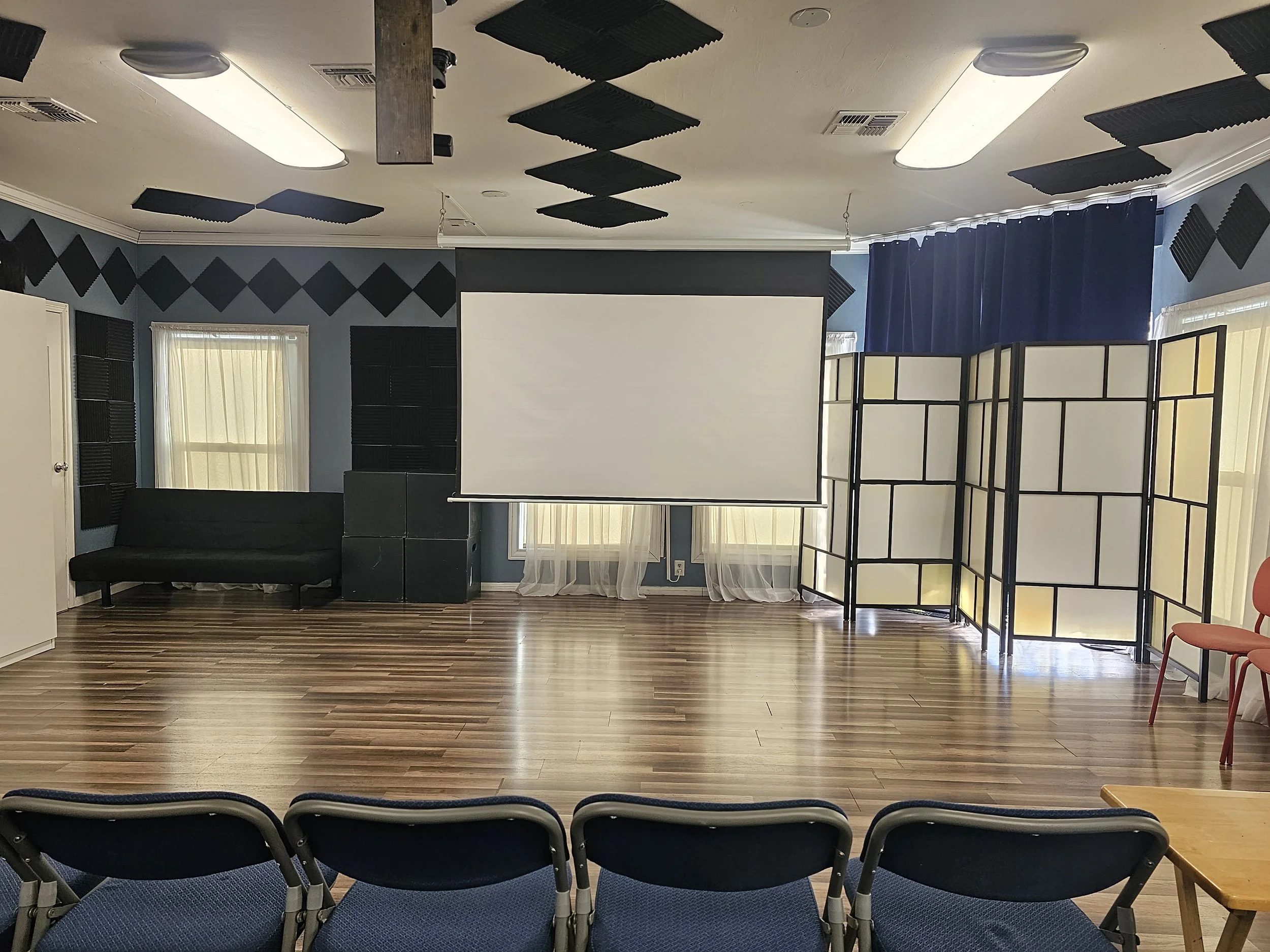 Studio E at Lounge on Melrose screening room with projector screen, black acoustic foam panels, couch, folding chairs, and room divider.