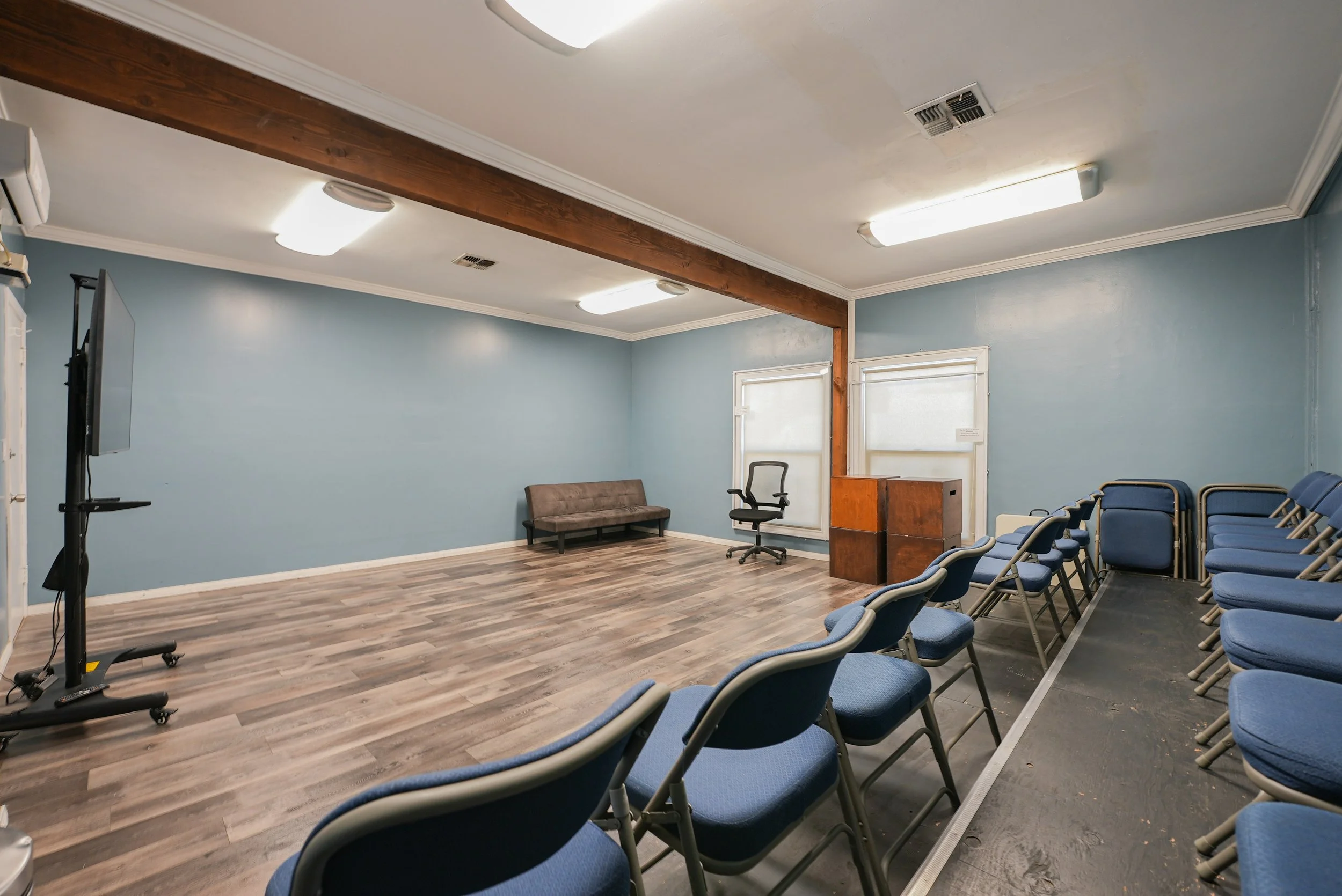 Studio C at Lounge on Melrose Empty blue-walled acting studio with blue walls, rows of blue chairs, a presentation screen, podium, couch, and wood-beam ceiling