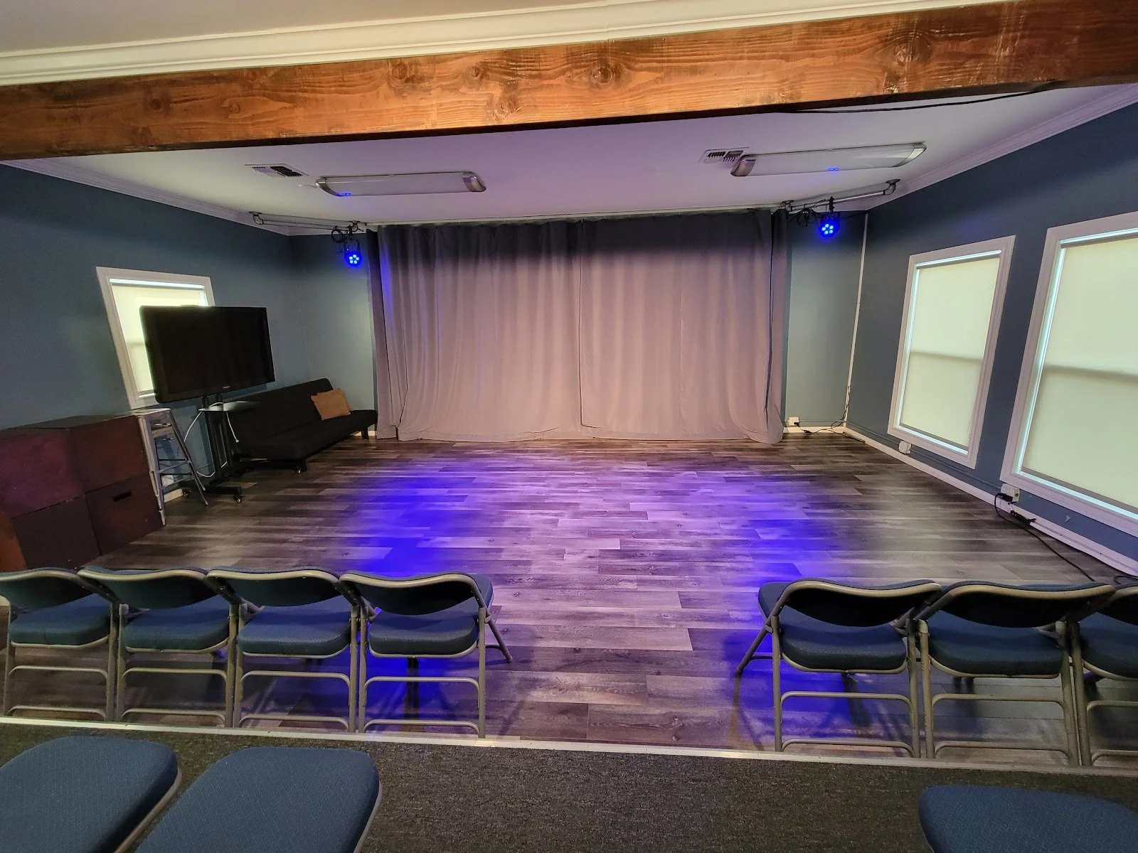 Studio D at Lounge on Melrose Small event venue with curtained stage, purple lighting, rows of folding chairs, a couch, and a television