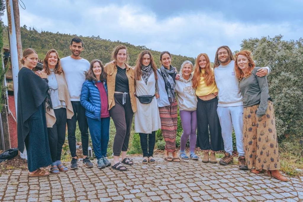 A group of people standing together outdoors on a stone path, with greenery and a cloudy sky in the background. They are smiling and dressed casually.
