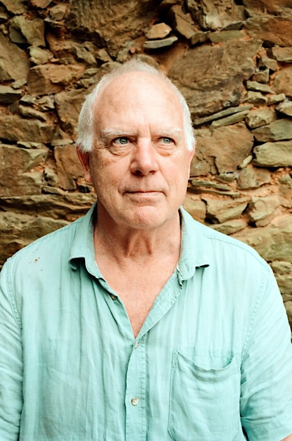 Elderly man with white hair and blue eyes wearing a light blue shirt standing against a stone wall.