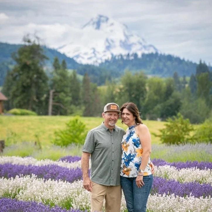 A smiling couple standing outdoors in a flower field with purple and white flowers, with Mount Hood in the background. Hope Ranch Lavender. Wedding, events, guests, Hood river, Oregon