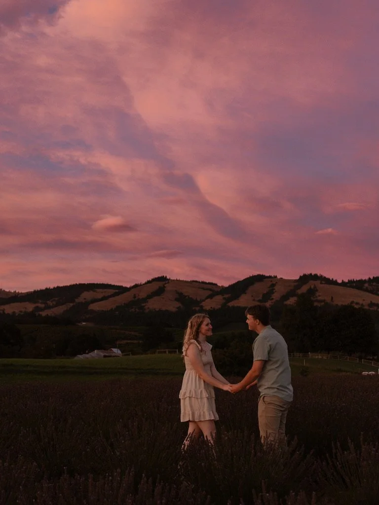 A couple holding hands in a lavender field during sunset with colorful pink and purple clouds and distant hills in the background. Lavender field in Hood River Oregon, engagement photo, Hope Ranch Lavender
