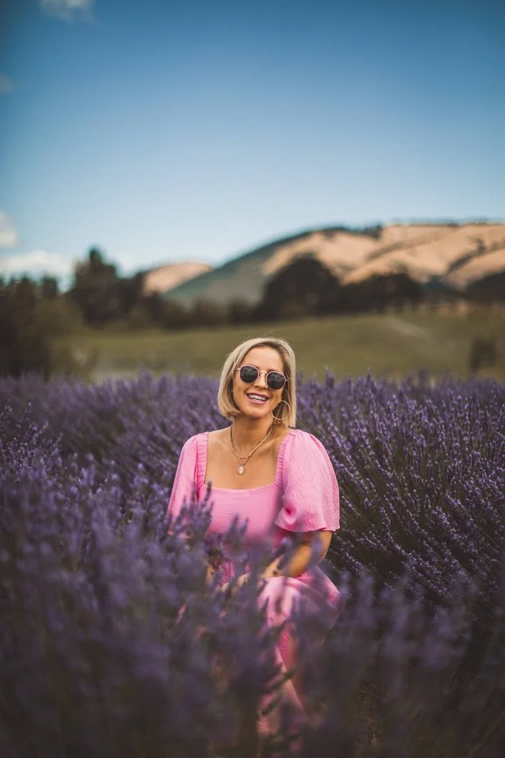 A woman sitting in a lavender field, wearing a pink dress and sunglasses, smiling with mountains in the background. lavender field photography, hope Ranch Lavender