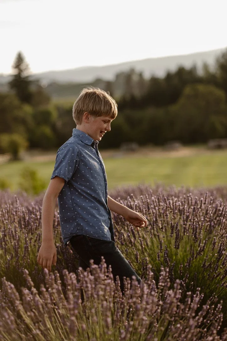 family photography in lavender field. A young boy with blonde hair wearing a blue short-sleeve button-up shirt with white polka dots and black pants walking through a lavender field during the daytime, smiling and looking down. Hope Ranch Lavender.