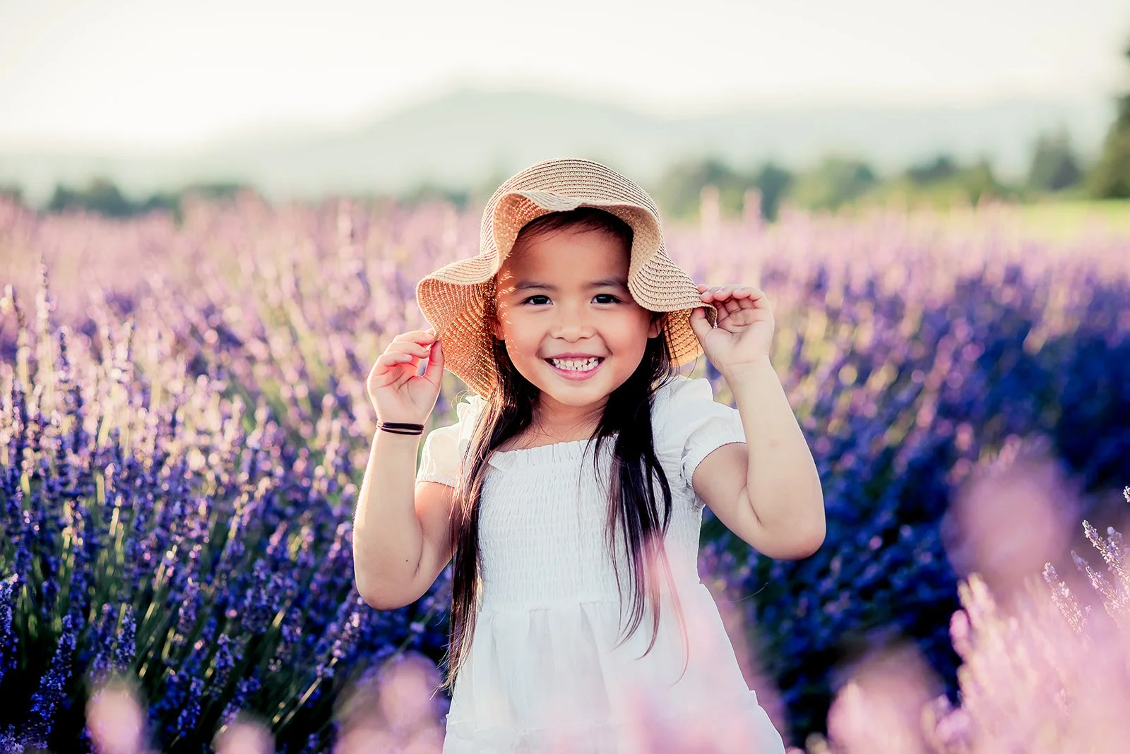 lavender field photography, A young girl with long dark hair wearing a white dress and a wide-brimmed straw hat, standing in a field of purple lavender flowers with mountains in the background, smiling and holding her hat. Hope Ranch Lavender