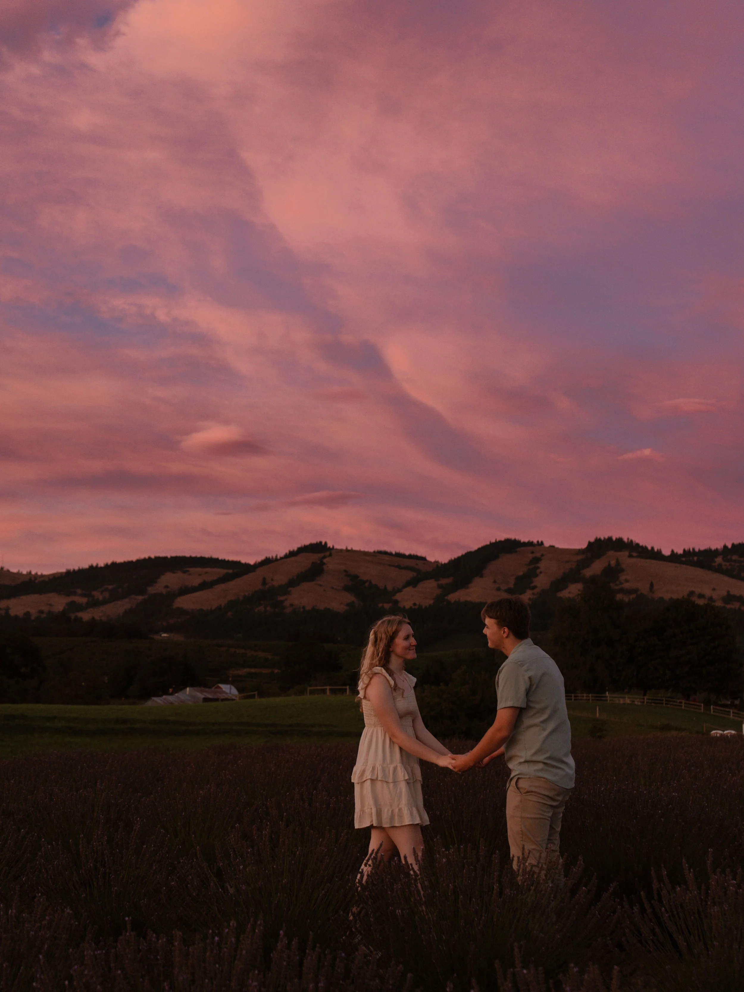 A couple holding hands and facing each other in a lavender field at sunset, with a colorful pink and purple sky and rolling hills in the background. Engagement photos Hood River