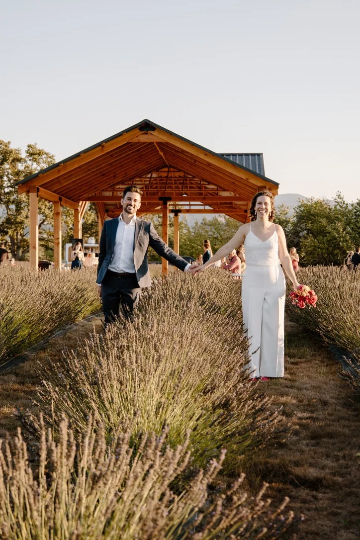 A couple in wedding attire holding hands and walking through a lavender field with a wooden pavilion in the background, smiling. Wedding venue Hood River, Oregon Hope Ranch Lavender