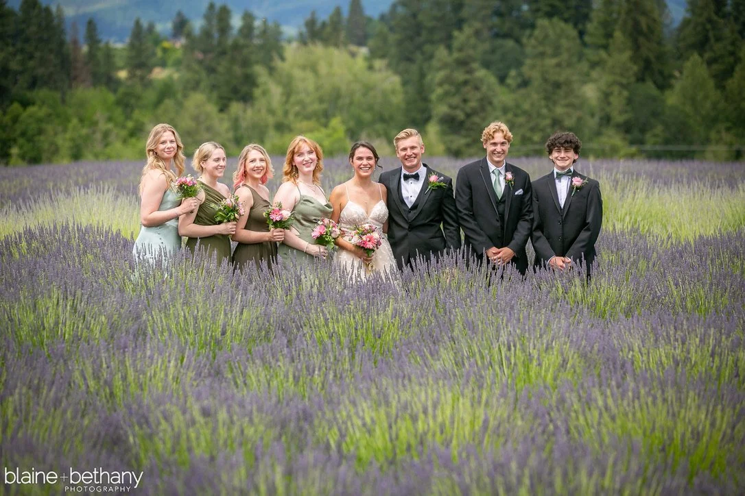 A wedding party standing in a lavender field with trees in the background, including a bride in a white dress, a groom in a tuxedo, and five women and men in formal dresses and suits, all smiling. Hope Ranch Lavender, Hood river, Oregon