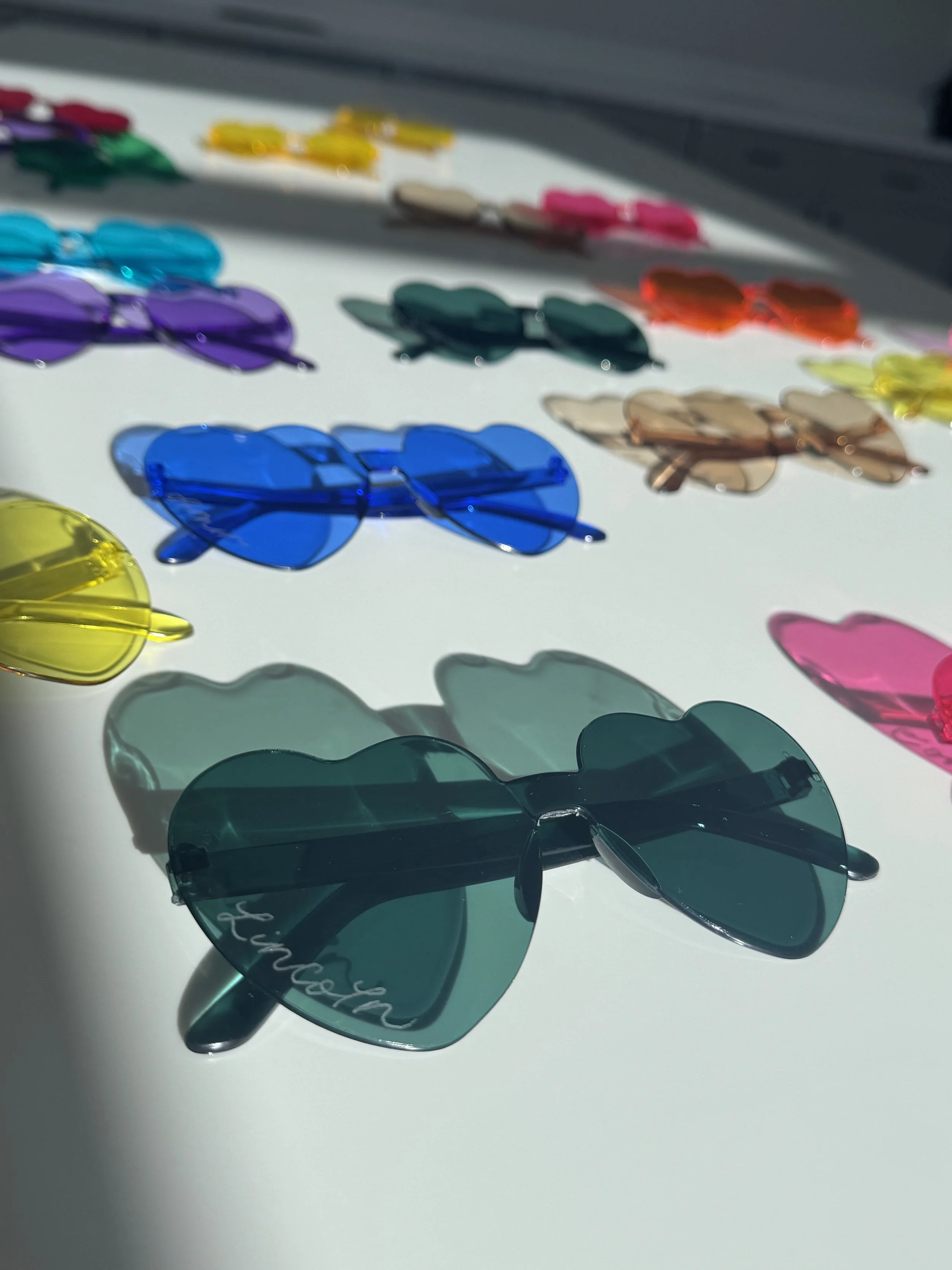 Colorful sunglasses arranged on a white surface with sunlight casting shadows.