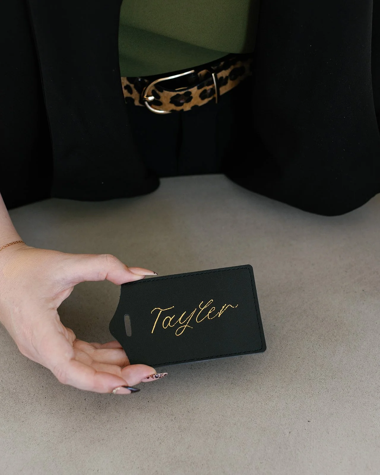 A hand holding a black leather luggage tag with gold embroidered name 'Tayler' in calligraphy, in front of a beige surface near a green and black outfit hanging with a leopard print belt.