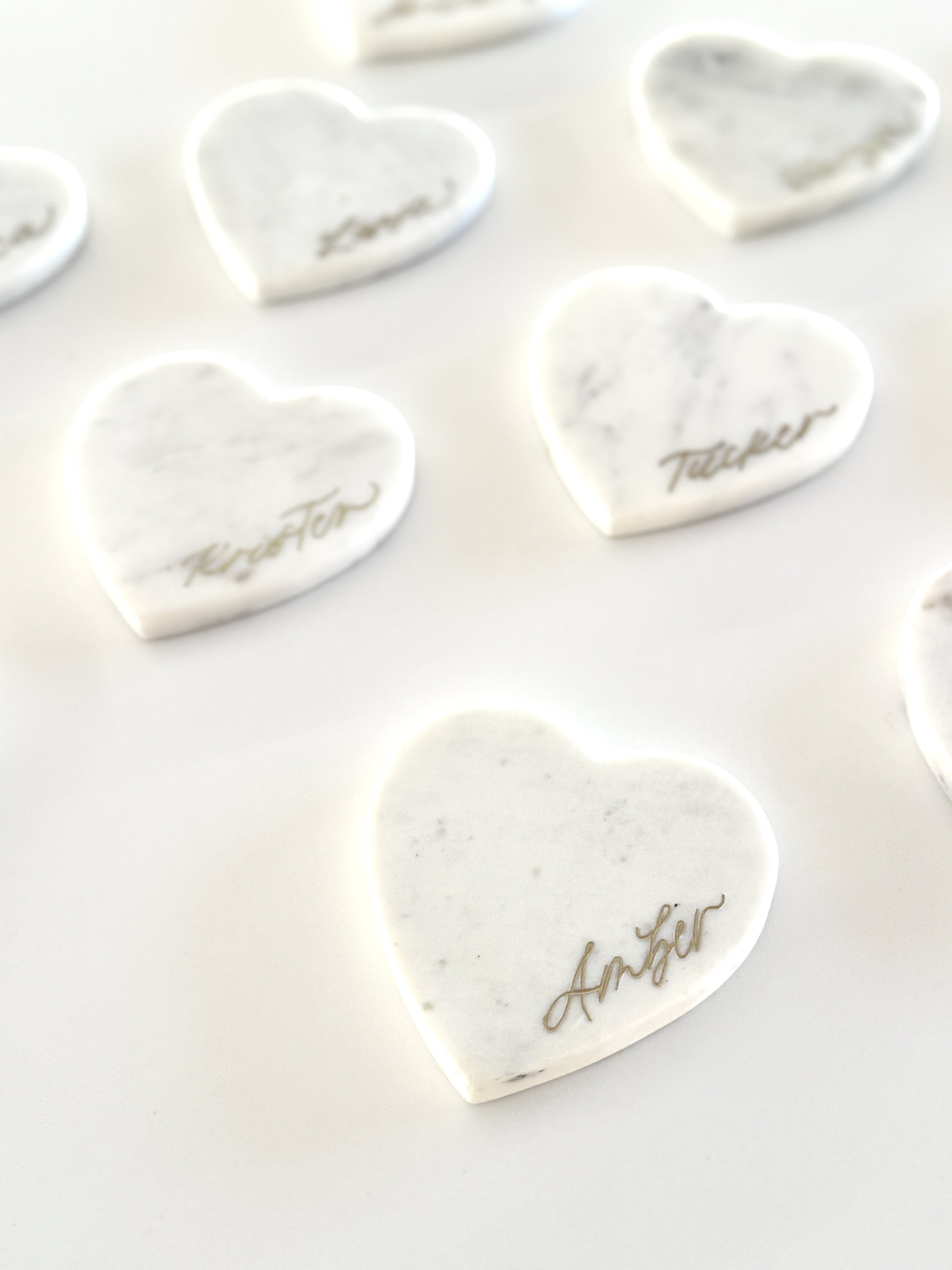 White marble heart-shaped tokens with handwritten words like "Love," "Forever," "Best," and "Amour" written in metallic script, arranged on a white surface.
