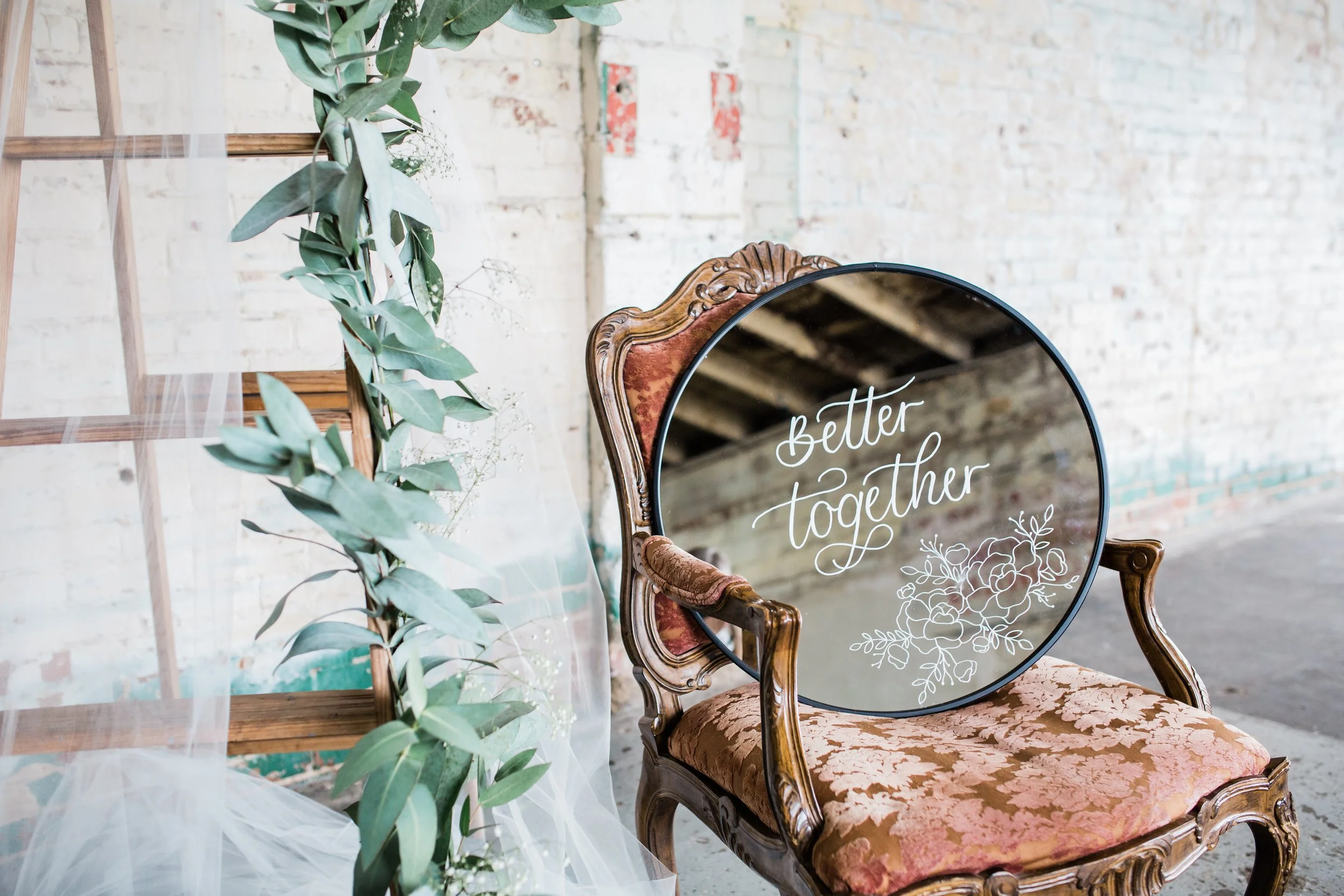 A round mirror with "Better Together" written in calligraphy sets on a vintage floral print chair.