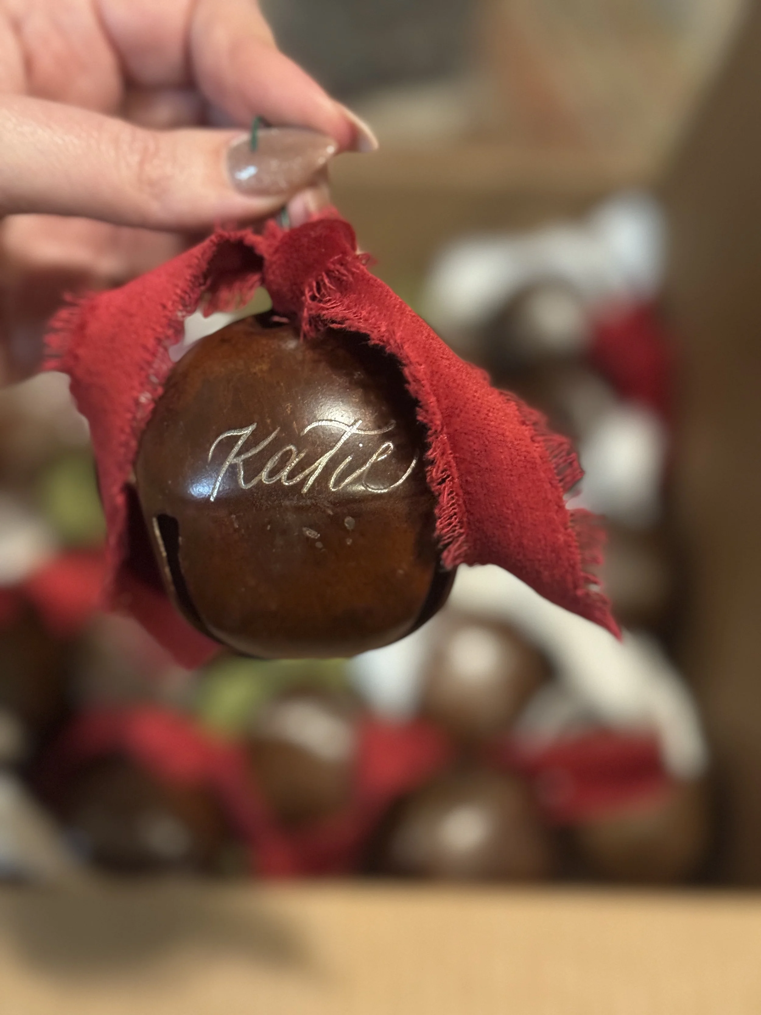A person holding a brown Christmas ornament with the name 'Kaitie' engraved on it, wrapped in a red cloth with frayed edges, with more similar ornaments blurred in the background.