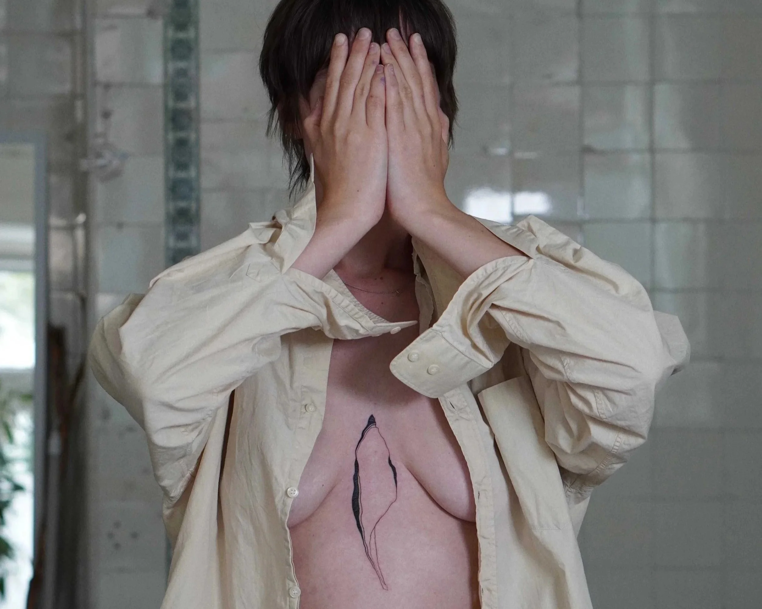 A person with short brown hair is covering their face with both hands. They are wearing a beige or cream-colored shirt, unbuttoned at the top, revealing a tattoo on their sternum. The background contains grayish tiles, suggesting an indoor setting.