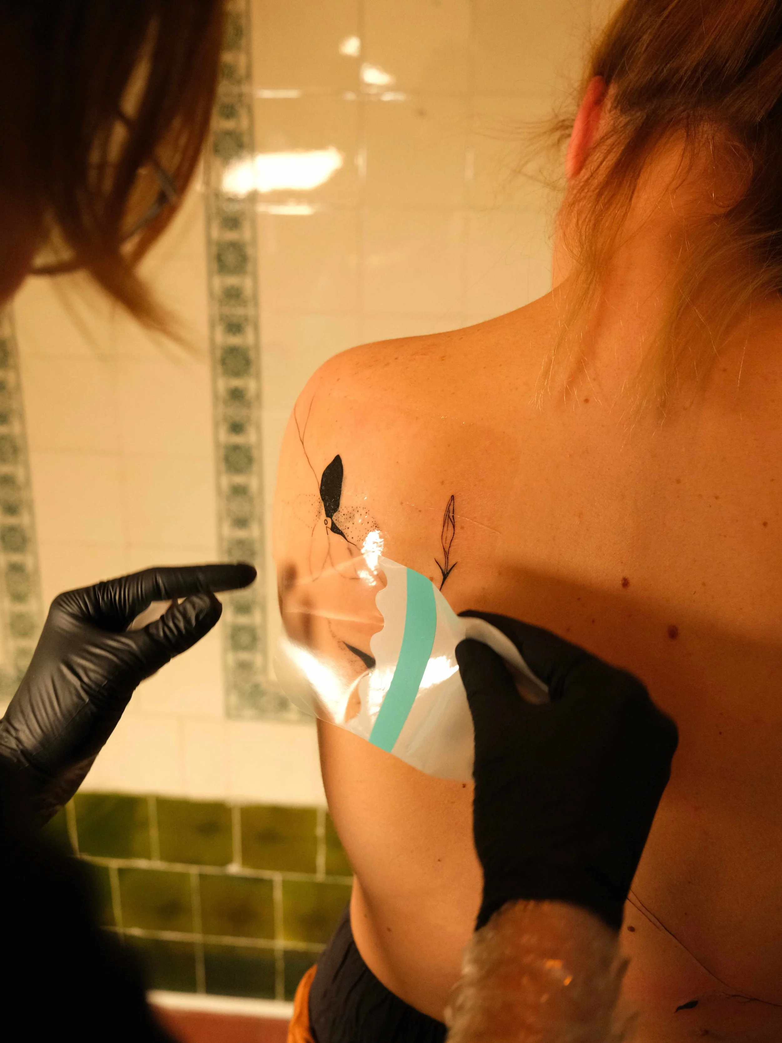 A person getting a tattoo on their shoulder in a tattoo studio, with a tattoo artist wearing black gloves holding a tattoo needle, and a tattoo design of a bird with branches being inked.