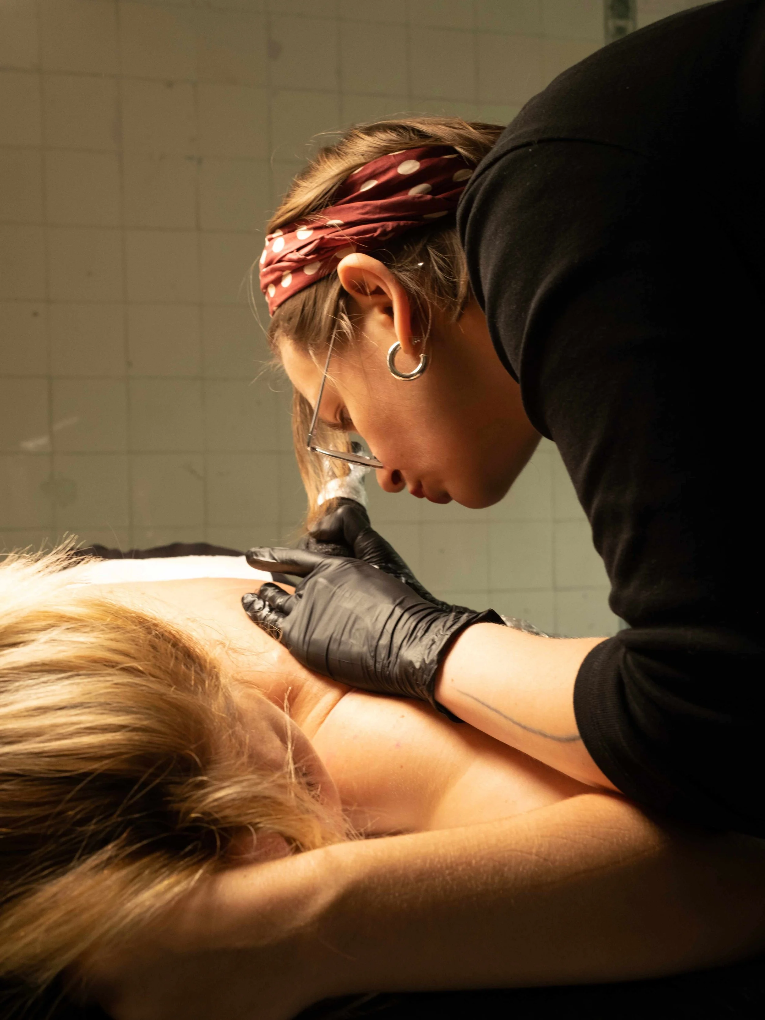 Anna Ustrehova, a tattoo artist wearing black gloves, glasses and earrings tattooing female client's back.