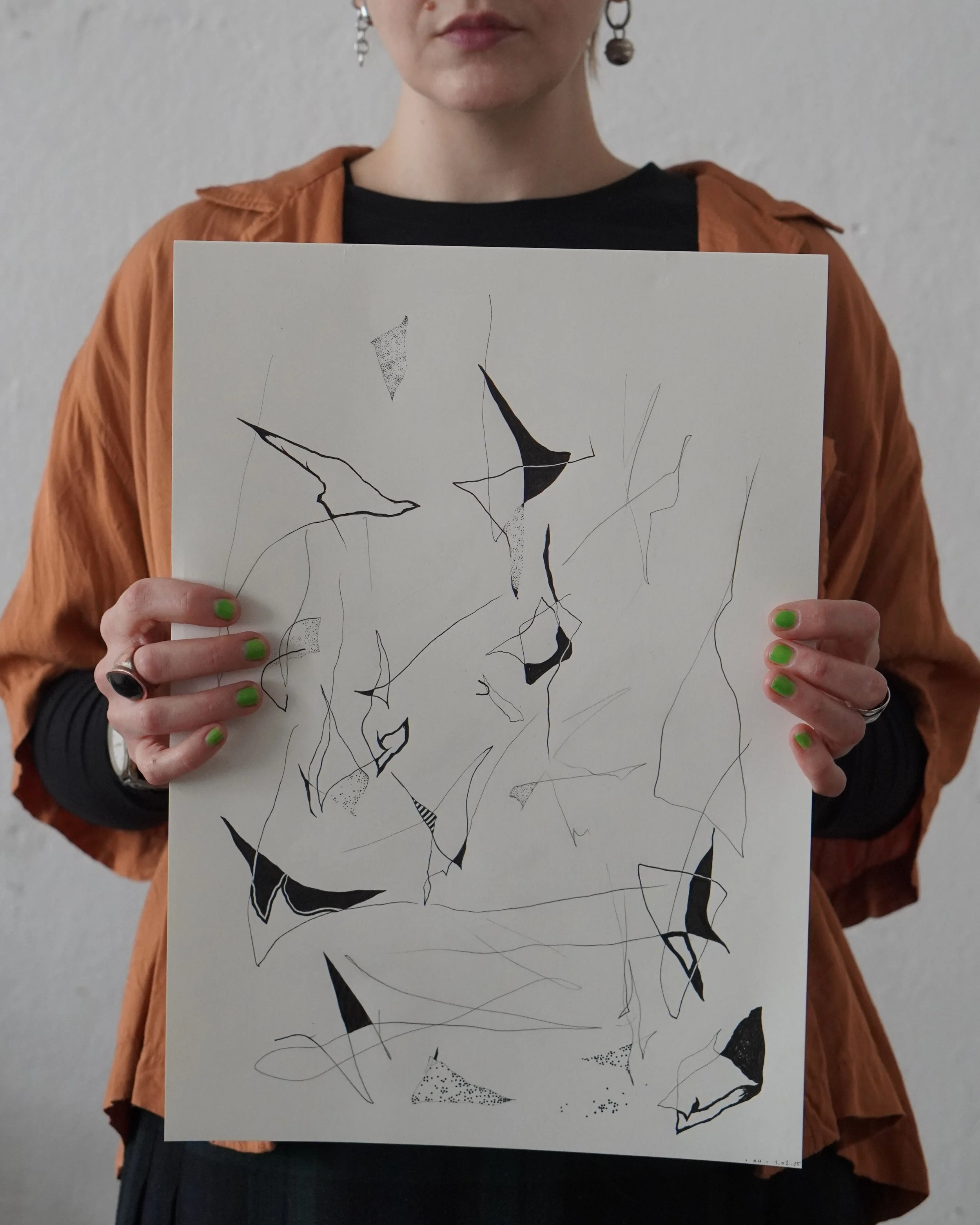 Artist holding a A3 original artwork with abstract black lines and shapes, wearing a black top and an orange jacket, with green nails and visible earrings, against a plain wall.