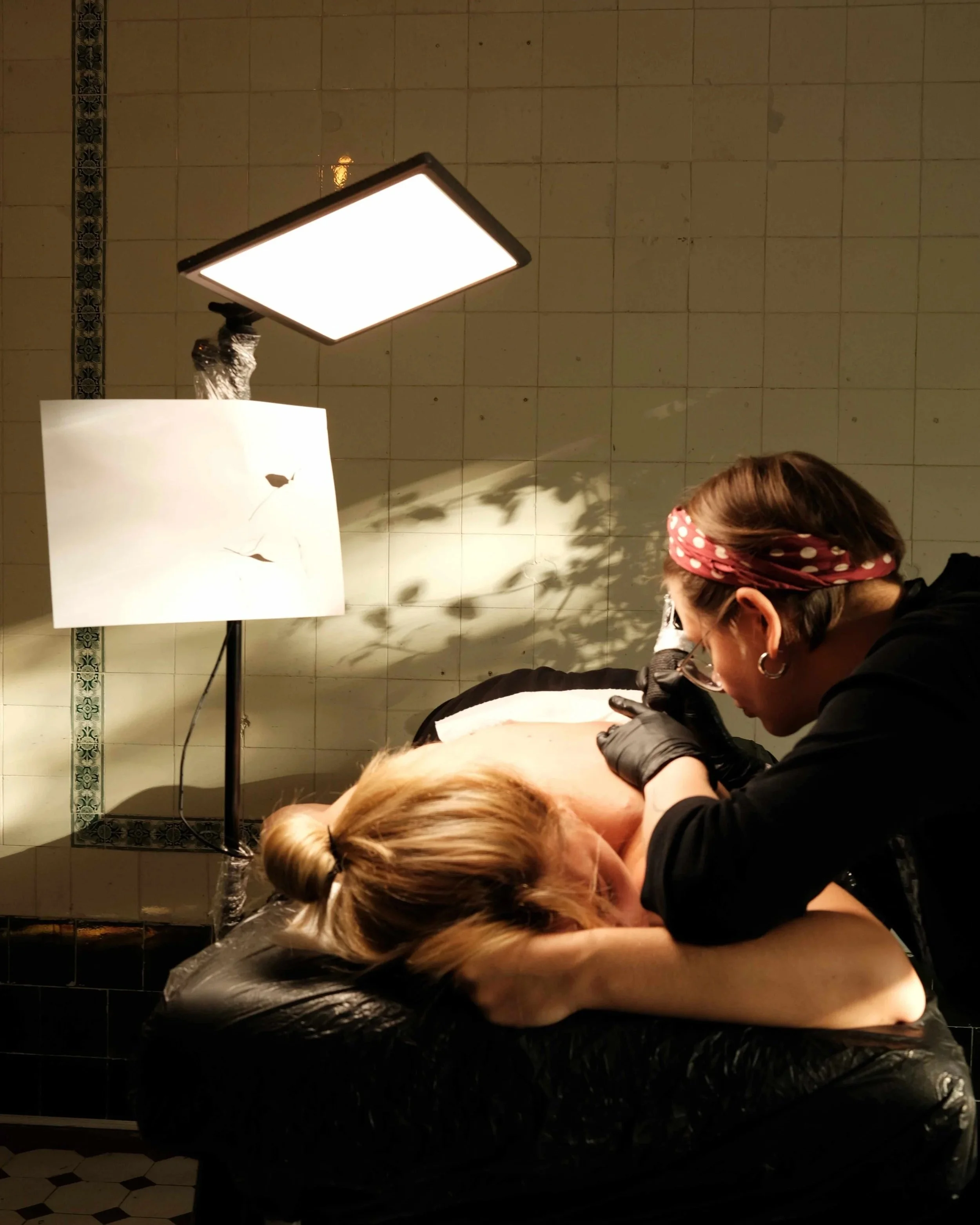 A woman getting a tattoo in a tattoo studio. The woman has blonde hair tied back and is lying on a tattoo table. The female tattoo artist, wearing black gloves, is working on her back tattoo. The background features tiled walls of Berlin Altbau.