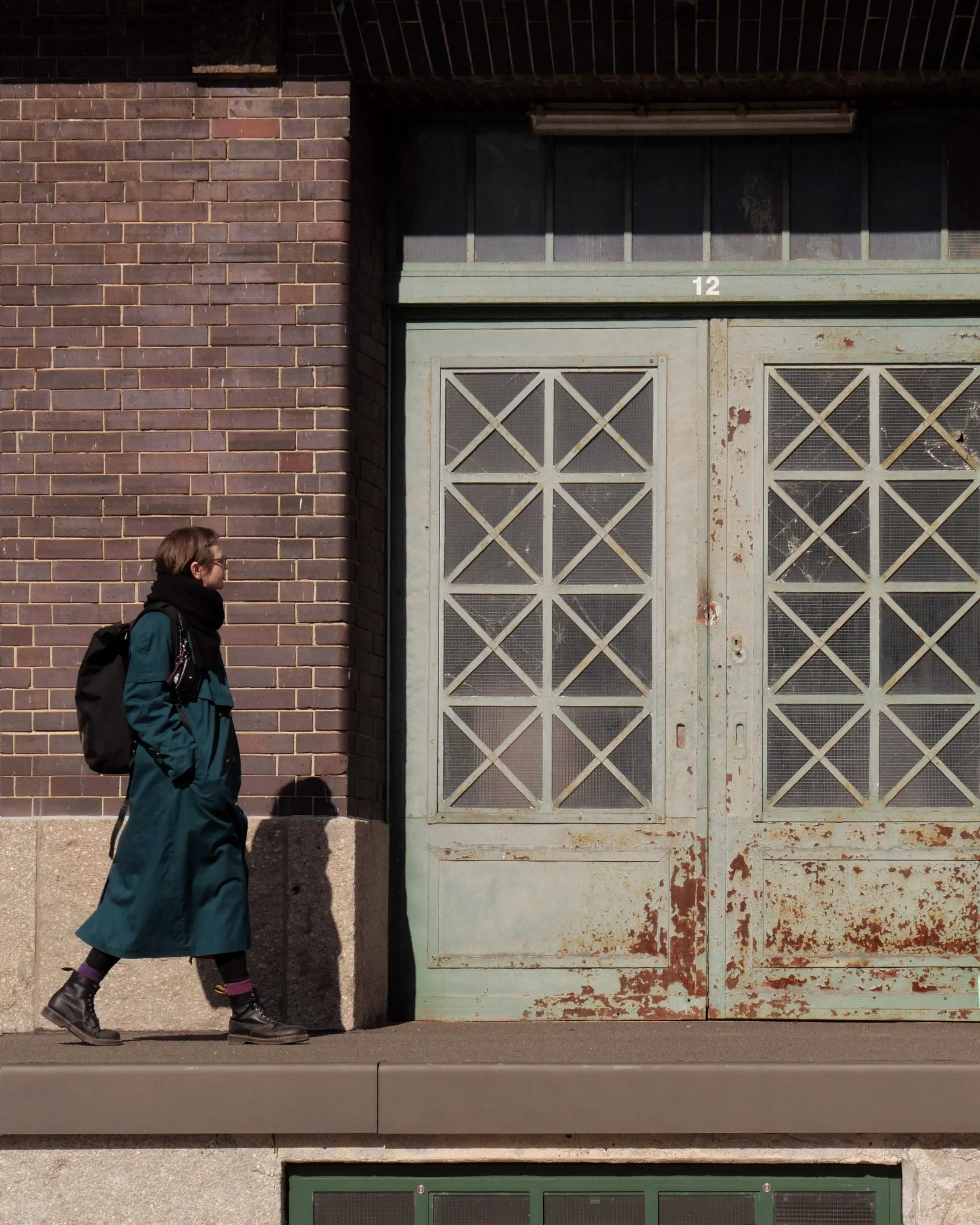 A woman with brown hair wearing glasses, a teal coat, a black scarf, black boots, purple socks, and carrying a black backpack walks past a large, weathered metal garage door with a geometric grid pattern. The garage door is mounted on a brick building with a small brick extension and a green window at the bottom.