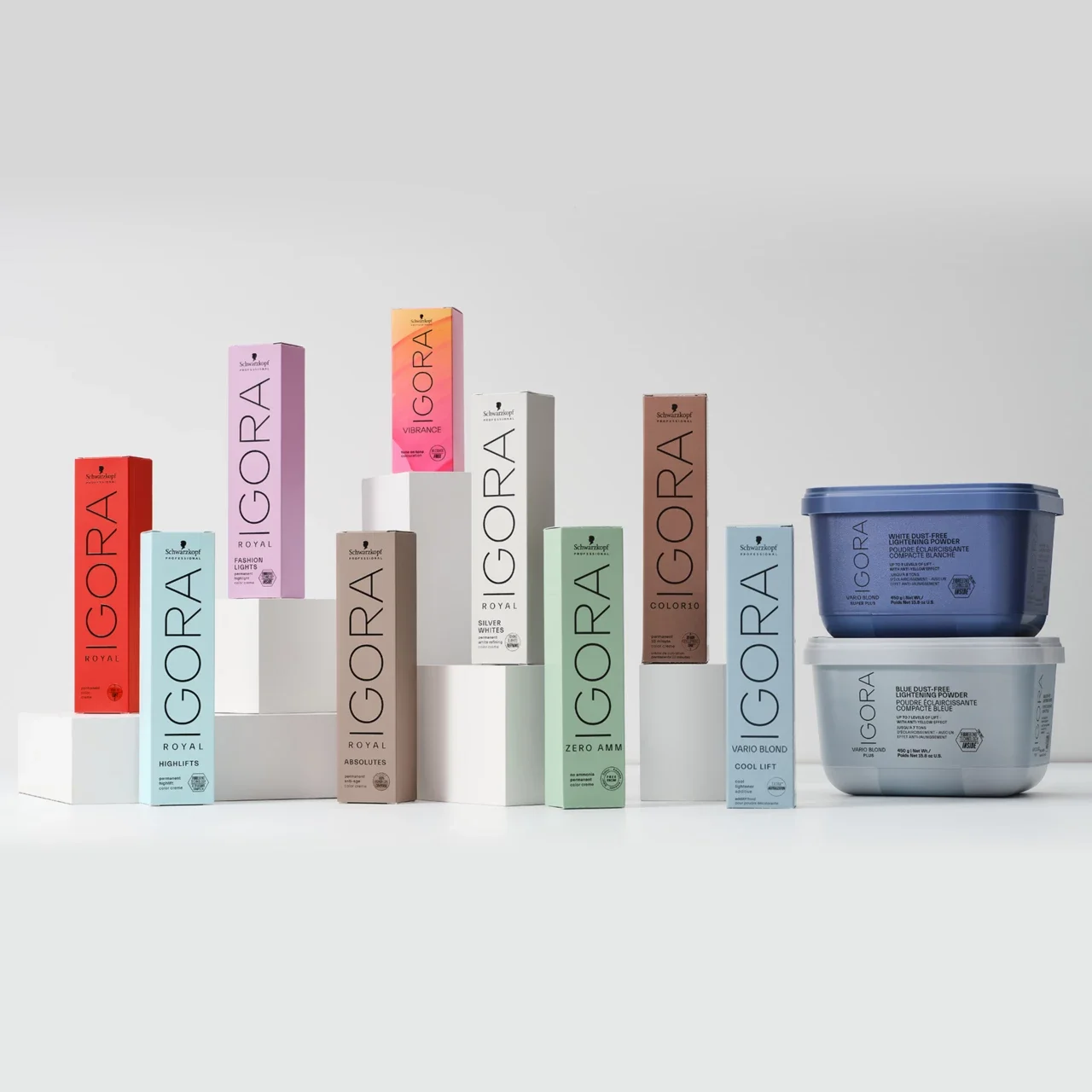 Collection of hair coloring products by Igora in various colors and two containers of lightening powder, arranged on white pedestals against a white background.
