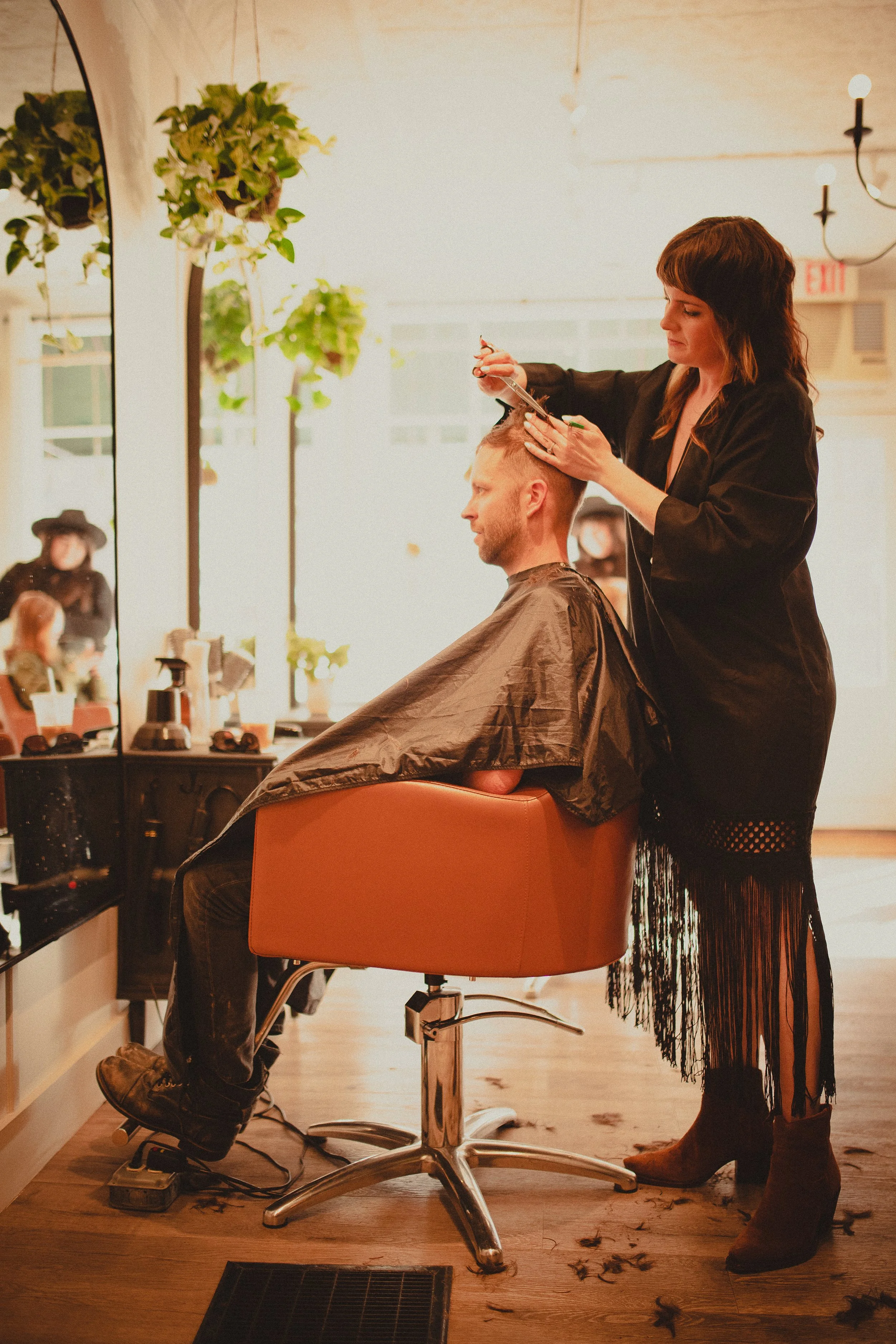 A hairstylist giving a haircut to a man sitting in a salon chair. The stylist is cleaning or styling the man's hair, who is wearing a brown cape. The salon has a large mirror, plants, and warm lighting, with hair on the floor.