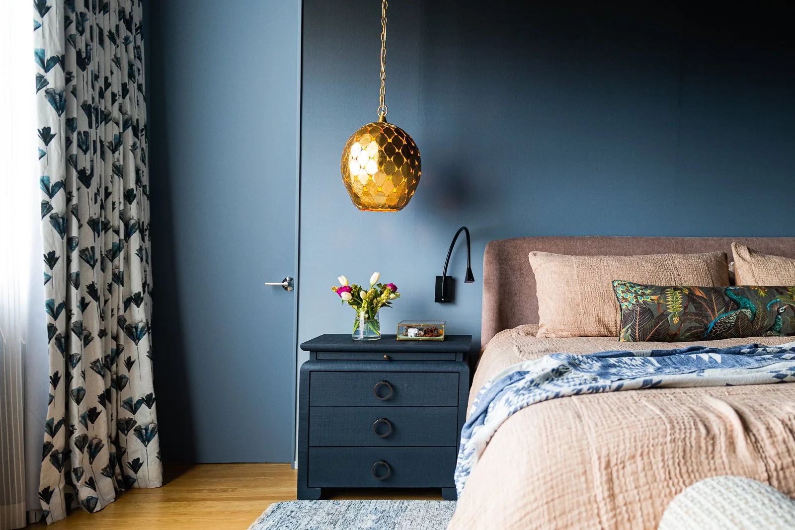 Primary bedroom nightstand styling with brass pendant lighting, dark blue walls, layered linen drapery, and minimalist reading lights