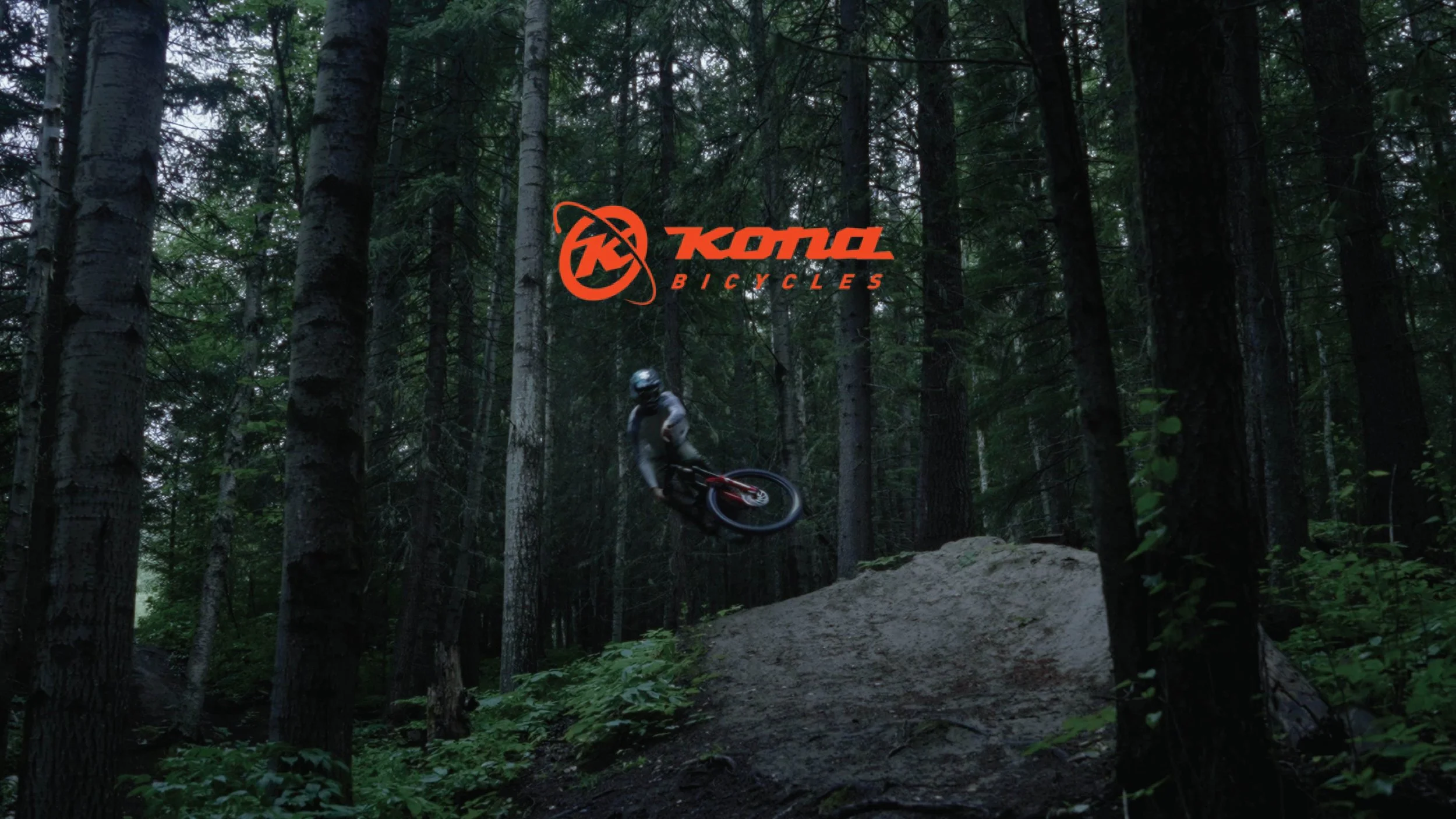 A mountain biker performing a jump on a dirt trail in a dense forest with tall trees and green foliage, with a Kona Bicycles logo overlay.