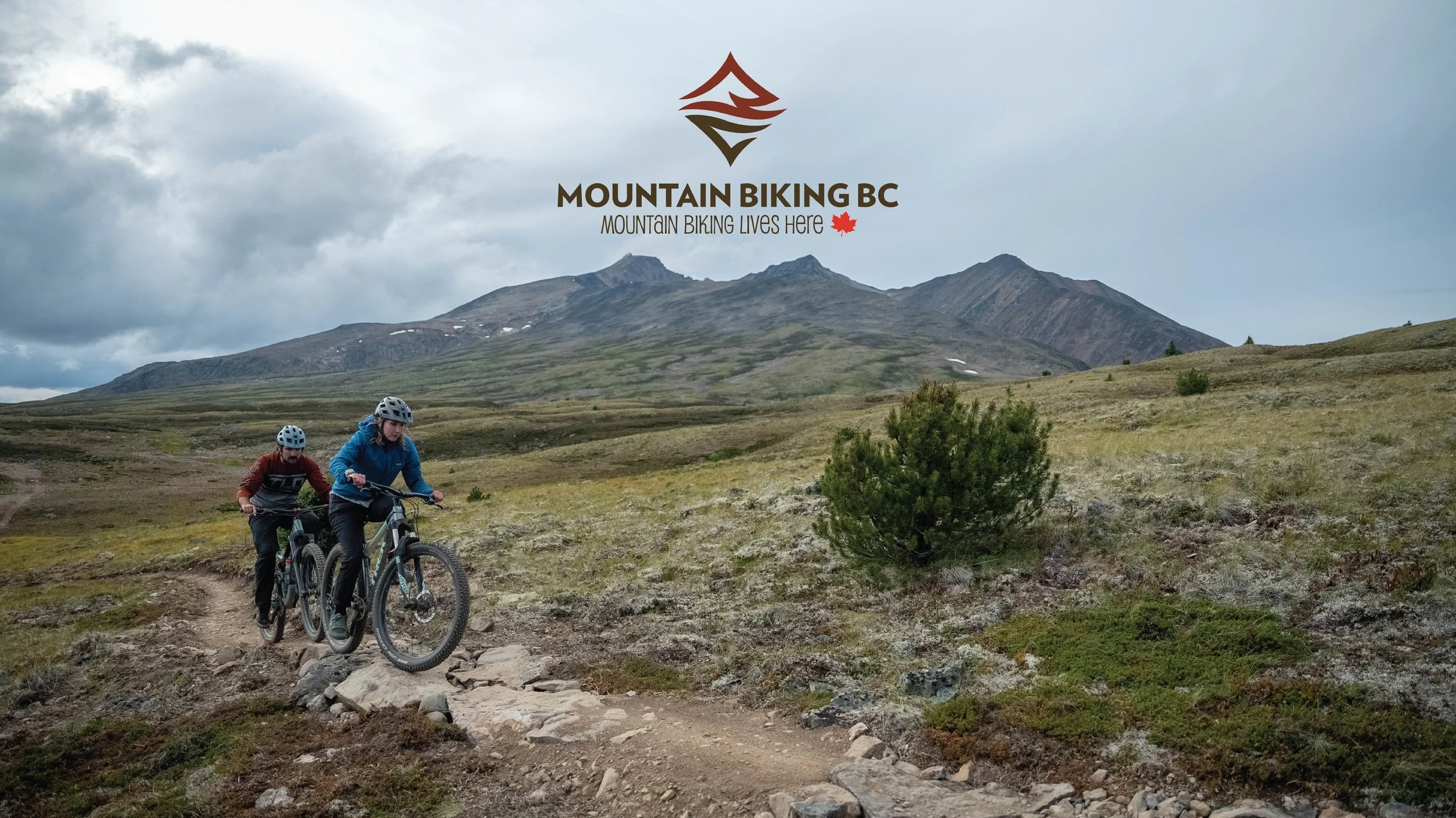 Two mountain bikers riding on a rocky trail in a mountainous landscape with green hills and cloudy sky, with a logo and text for Mountain Biking BC at the top.