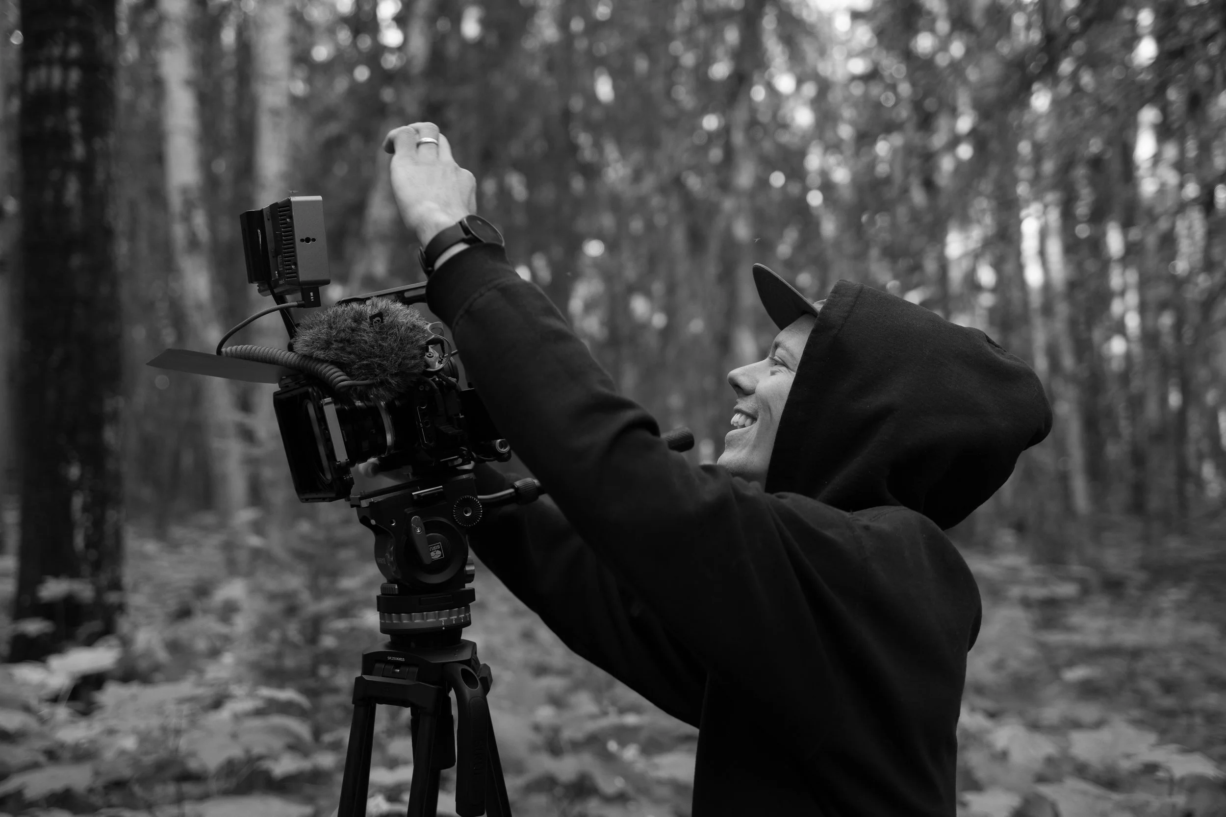 A person in a hooded jacket and hat operating a camera on a tripod in a forest with blurred trees in the background.