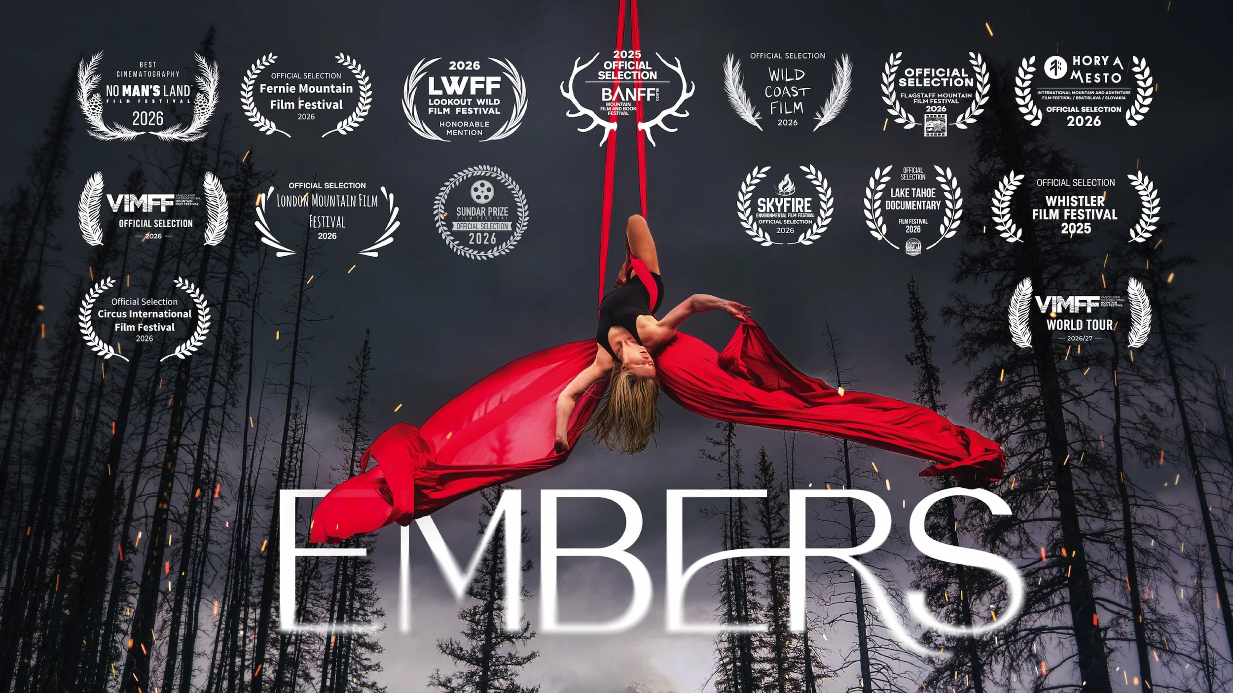A woman in a black top and red flowing skirt suspended upside down from red aerial silks amid a dark, cloudy forest background with tall, bare trees and fire sparks.