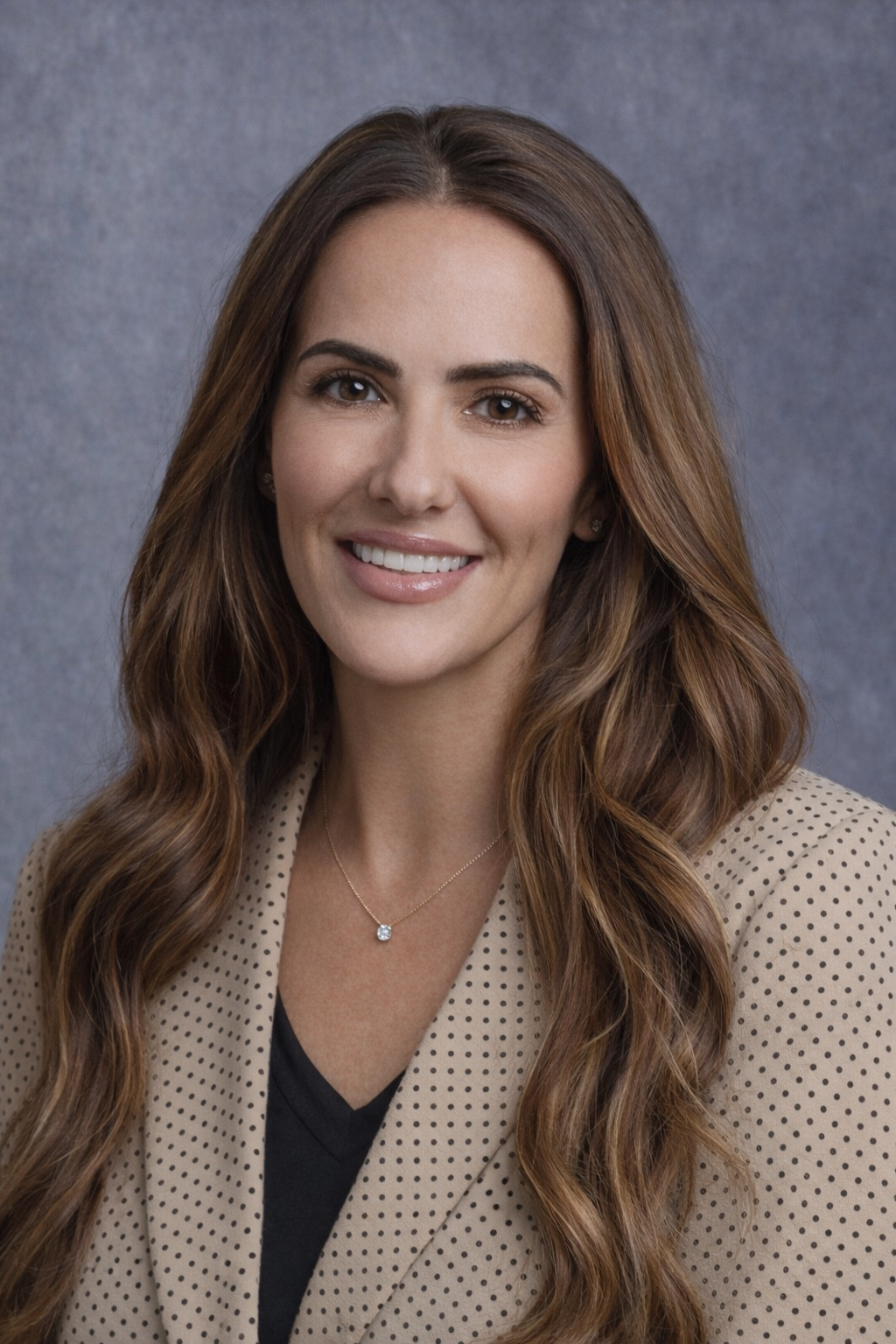 A woman with long wavy brown hair wearing a black long-sleeve top and gold jewelry stands next to a hospital bed covered with a white blanket in a modern room with dark textured wall panels.