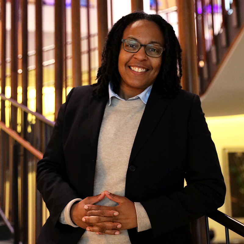 A smiling woman wearing glasses and a blazer over a sweater and collared shirt leans agains a stair rail
