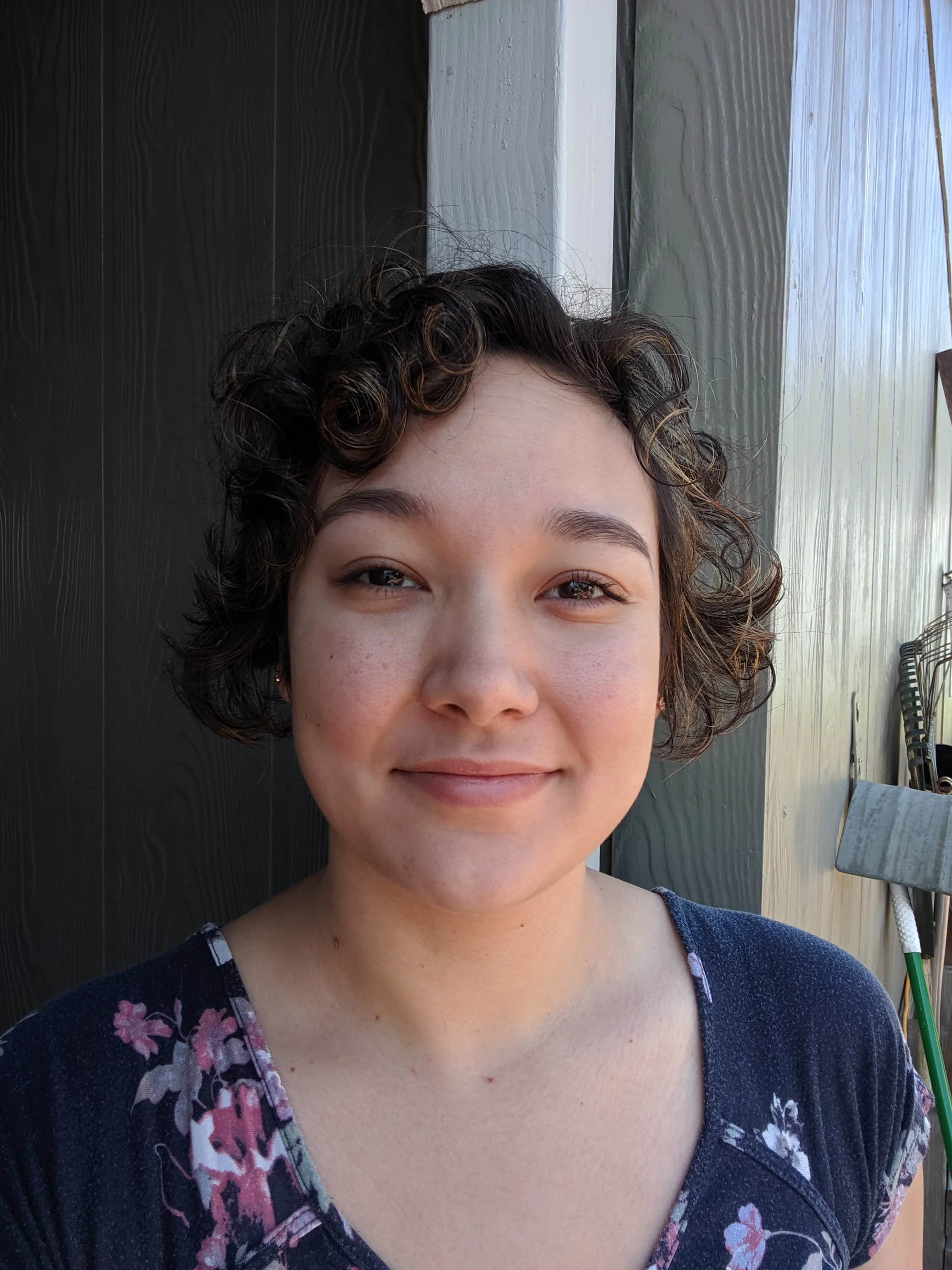 A young woman with short curly brown hair and light skin smiling outdoors with a gray wooden wall behind her, wearing a navy blue top with floral patterns.
