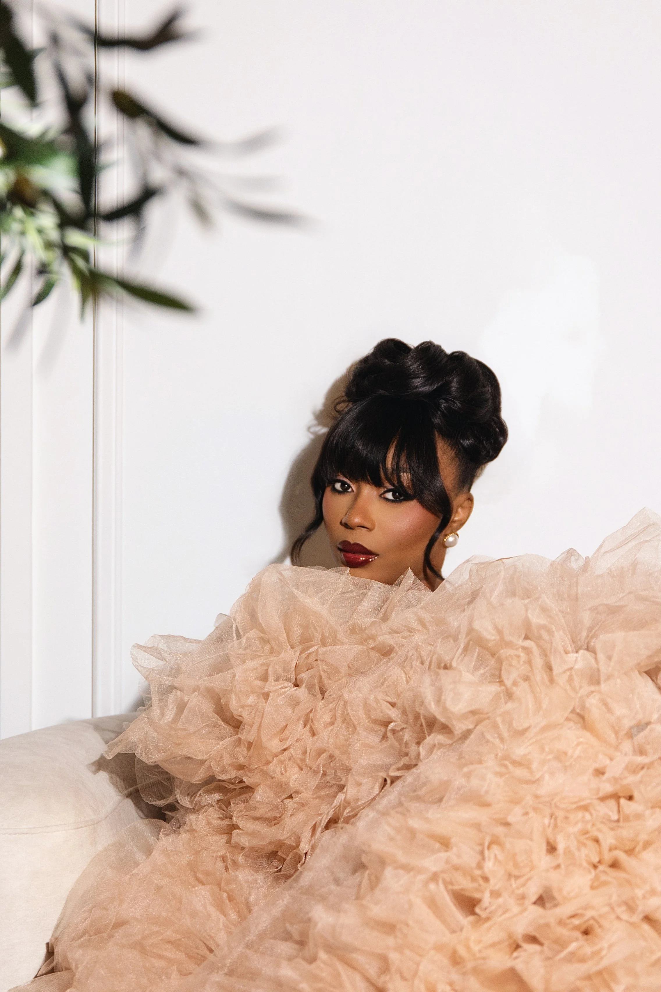 A woman with dark hair styled in an elegant updo, wearing makeup and pearl earrings, is partially covered with a voluminous, ruffled beige dress and is sitting against a plain white wall.