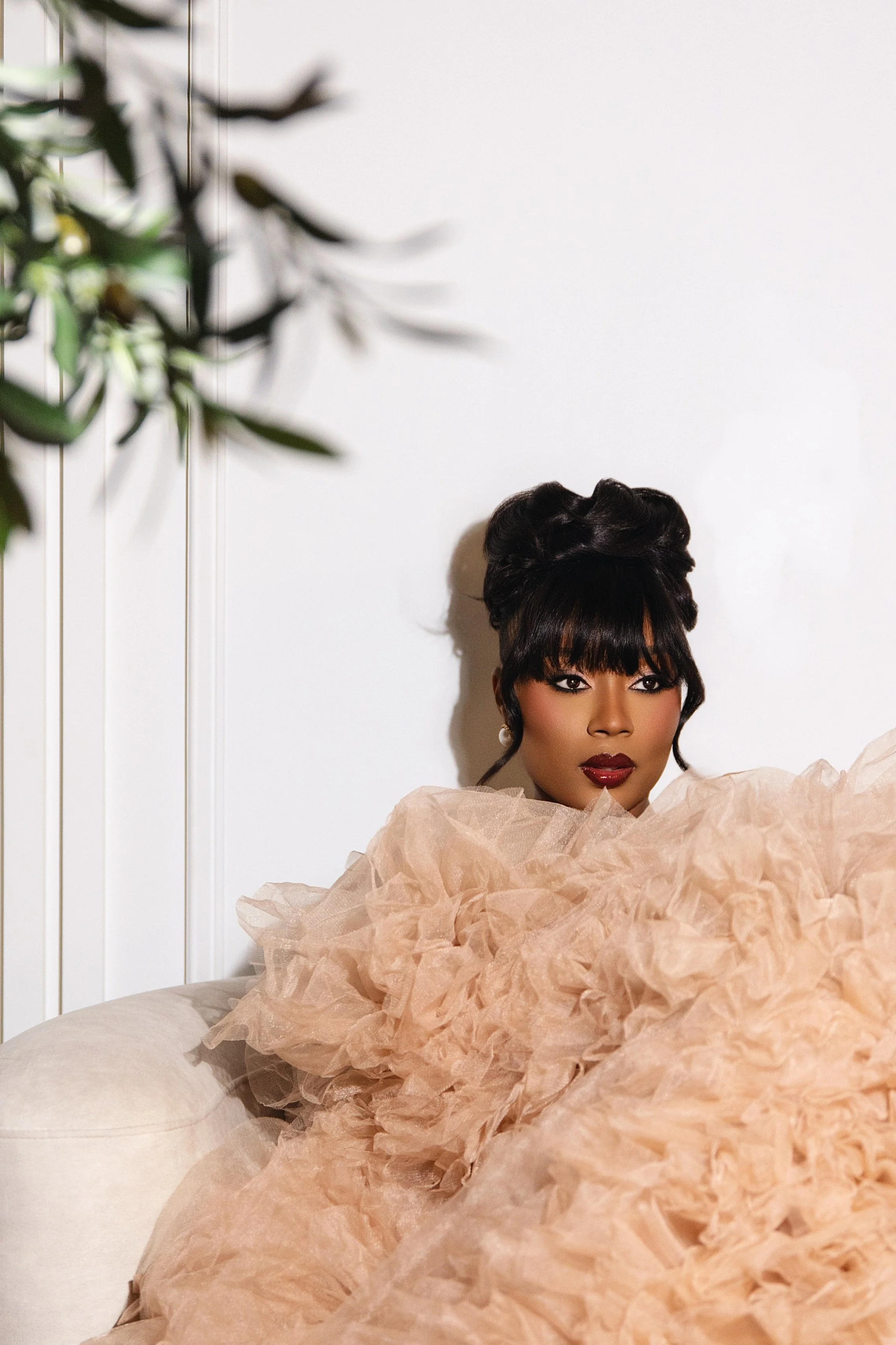 A woman with dark hair styled in an updo with bangs, wearing makeup with dark lipstick and eye makeup, is sitting on a beige sofa against a white wall. She is wearing a voluminous, ruffled peach-colored gown that covers her shoulders and part of her 