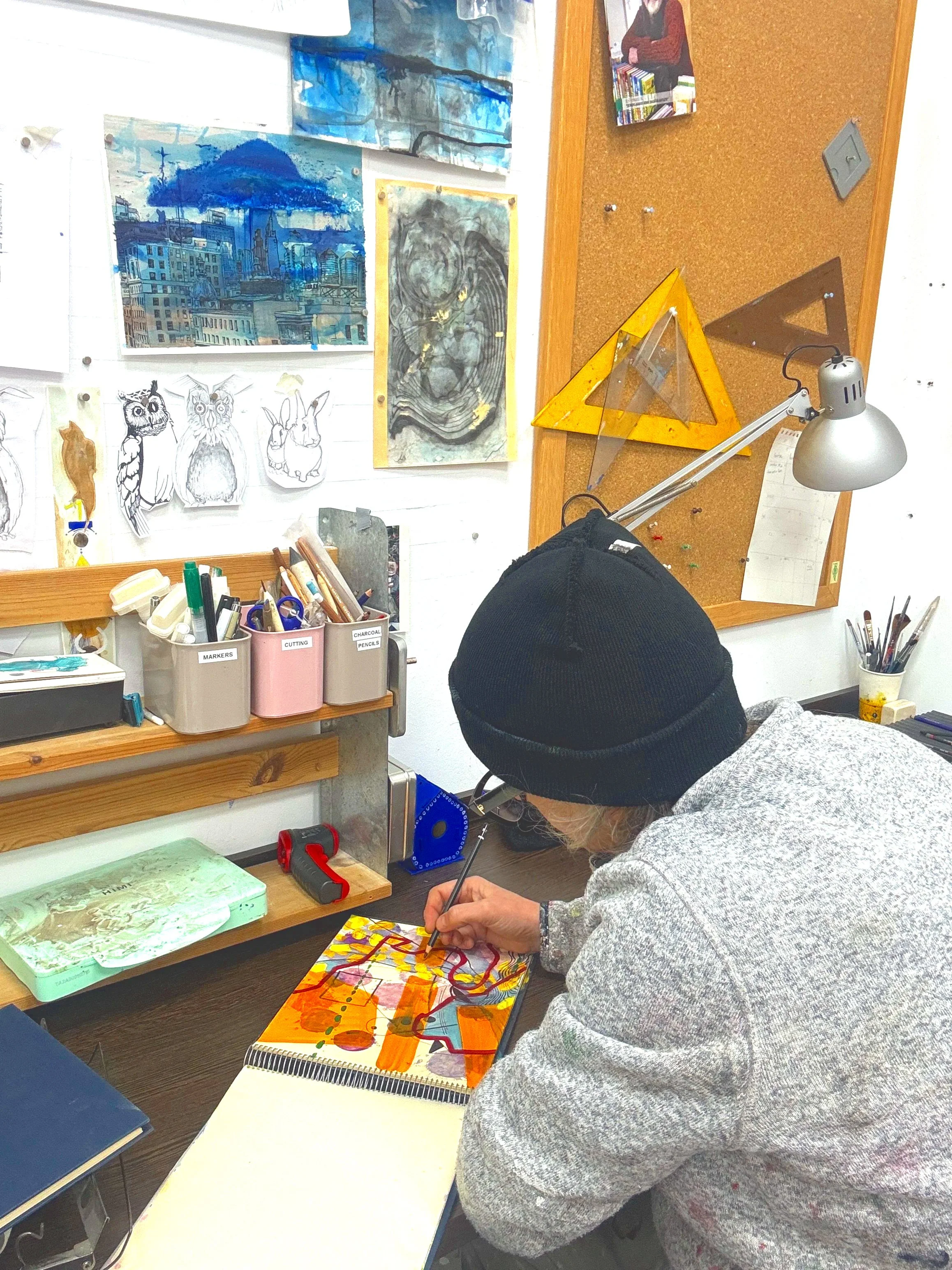 A person wearing a black beanie and gray hoodie working on a colorful abstract painting in an art studio with various paintings, drawings, and art supplies visible around.