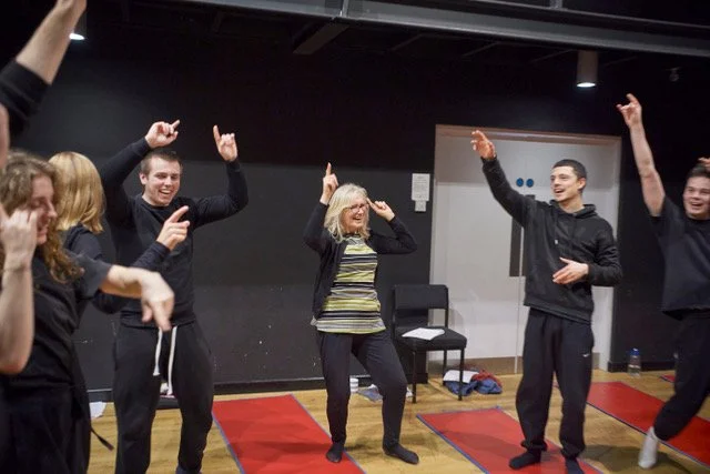 Group of people dancing and having fun in a room with a black wall and red yoga mats on the floor.