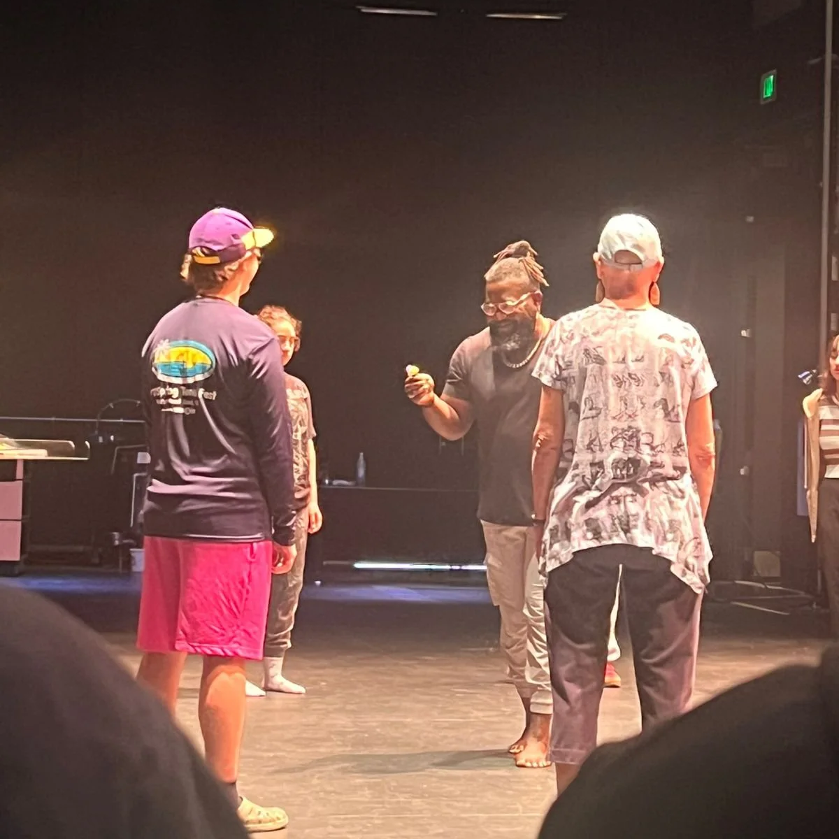Group of five people standing on stage, engaged in conversation. One person is holding an object, possibly a small dessert or snack. The background is dark with stage equipment visible.