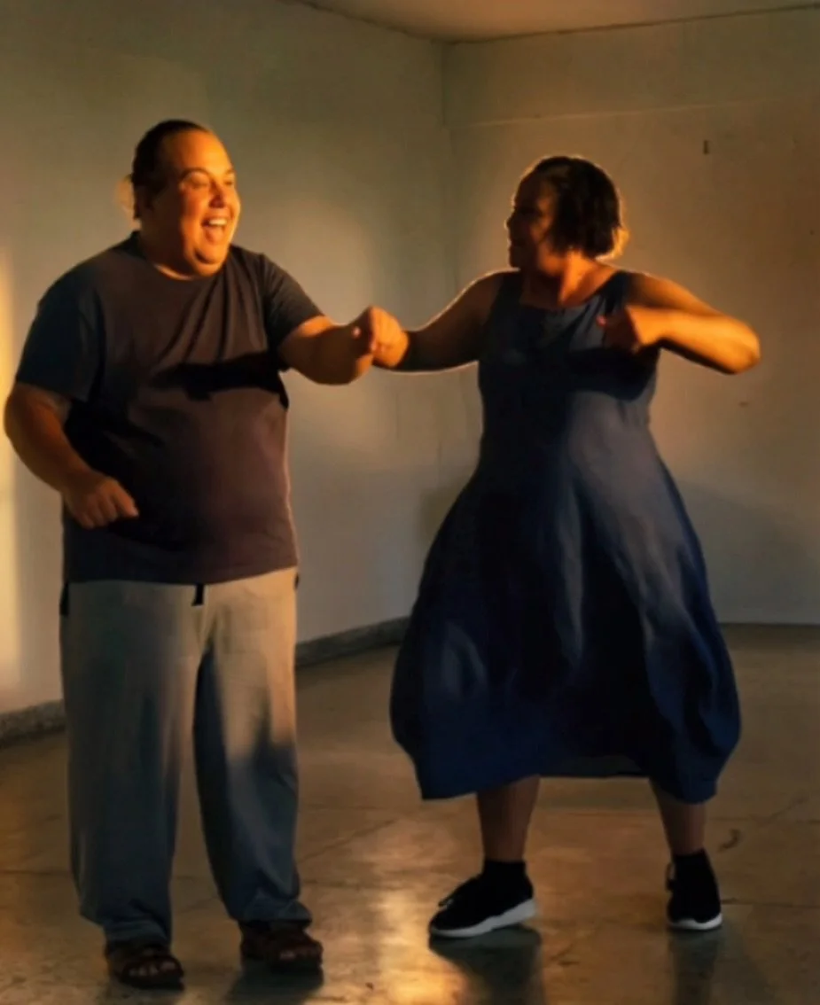 Two women are dancing and holding hands, one is wearing a dark t-shirt and light-colored pants, the other is wearing a dark dress and sneakers, in a dimly lit room.