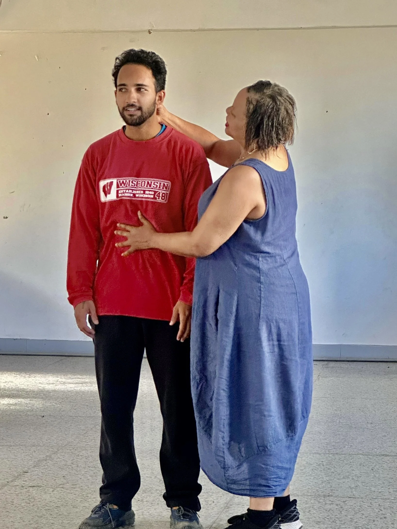 A woman is touching a young man's neck from behind while he is standing in front of her. The young man is wearing a red sweatshirt with Wisconsin and a number on it, and the woman is dressed in a blue dress.